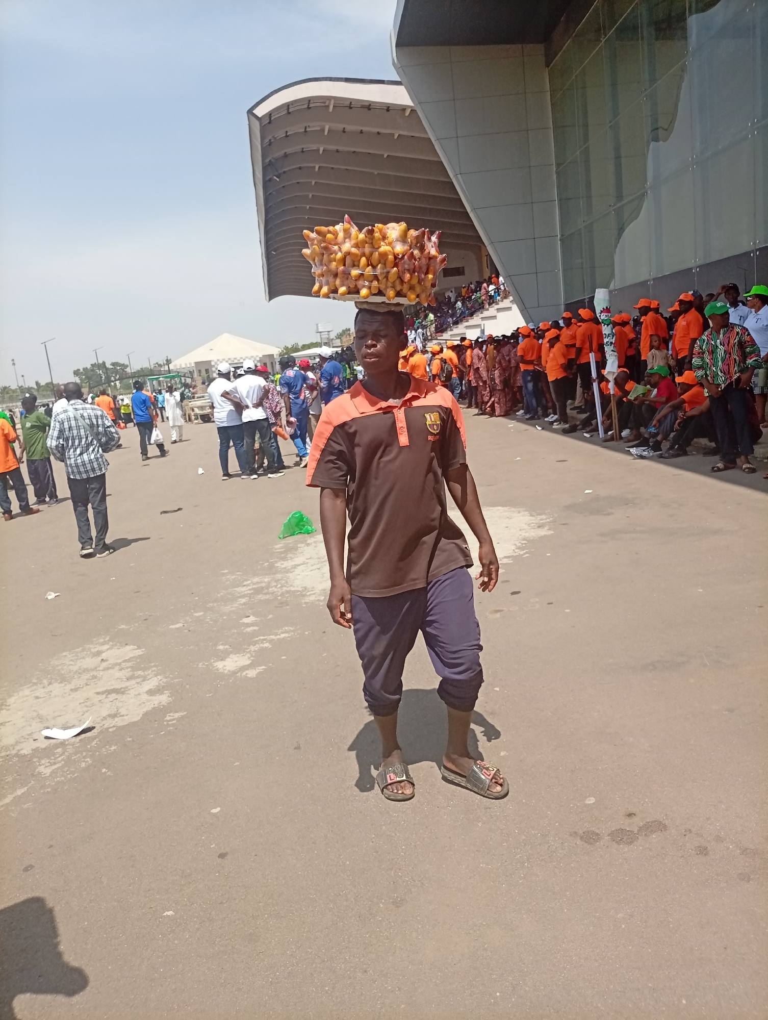 The image captures an outdoor scene in Unguwar Sarki, Nigeria, featuring a young man balancing a tray of individually packaged corn on his head, walking on a paved surface. He is the central figure, wearing a two-toned brown and orange polo shirt, dark blue trousers, and open-toed sandals. A small, blurry emblem is visible on his shirt, resembling a sports club logo.
Behind the vendor, a large crowd of people is gathered. Many individuals in the background are dressed in bright orange shirts and caps, suggesting a uniform or affiliation for an event. Some people wear green caps or checkered shirts, while others are in various casual attire. A few members of the crowd are seated along the base of a large, modern building.
The setting is characterized by a wide, paved area, likely a road or plaza, leading up to a distinctive contemporary building with a curved, cantilevered roofline and extensive reflective glass panels on its façade, typical of a stadium or large public venue. In the mid-ground, a white tent is visible. The ground shows some scattered litter, including plastic bags.
The activity depicted is that of street vending, with the young man hawking corn to the assembled crowd. The crowd itself appears to be congregated, possibly waiting for or attending an event, given the uniform clothing of many attendees.
Notable details include the bright, clear sky and strong sunlight, indicating midday, with distinct shadows cast on the ground. The architectural style of the large building contrasts with the informal gathering. Among the orange-clad crowd, a white sign or banner is partially visible, with the word "OFFICIAL" and a green logo being somewhat decipherable. The branding on the vendor's slippers, "PO LOL," is also visible.