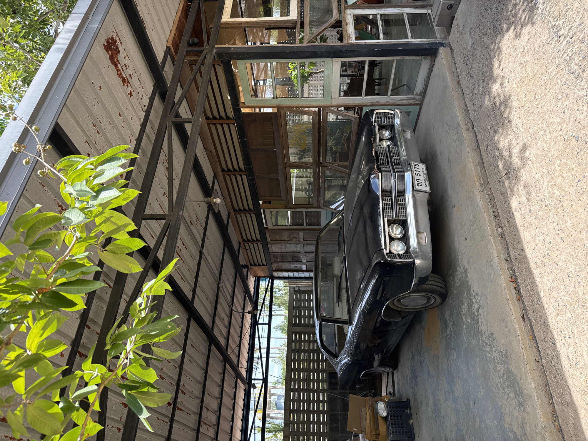 A vintage black sedan is parked inside an open-air structure with a corrugated metal roof supported by metal beams. The car is positioned on a concrete floor, facing towards the right of the frame. In front of the car, a license plate is visible. The structure appears to be an outdoor area, possibly a workshop or a covered parking space, with wooden walls and several windows on the right side. Behind the car, a concrete block wall with horizontal openings is visible, and beyond that, trees and a blue sky can be seen. To the left of the car, stacked boxes and a black crate are present. The overall environment suggests a rustic or industrial setting in a warm climate, consistent with a location in the Ko Samui District of Thailand. The image is well-lit by natural sunlight.