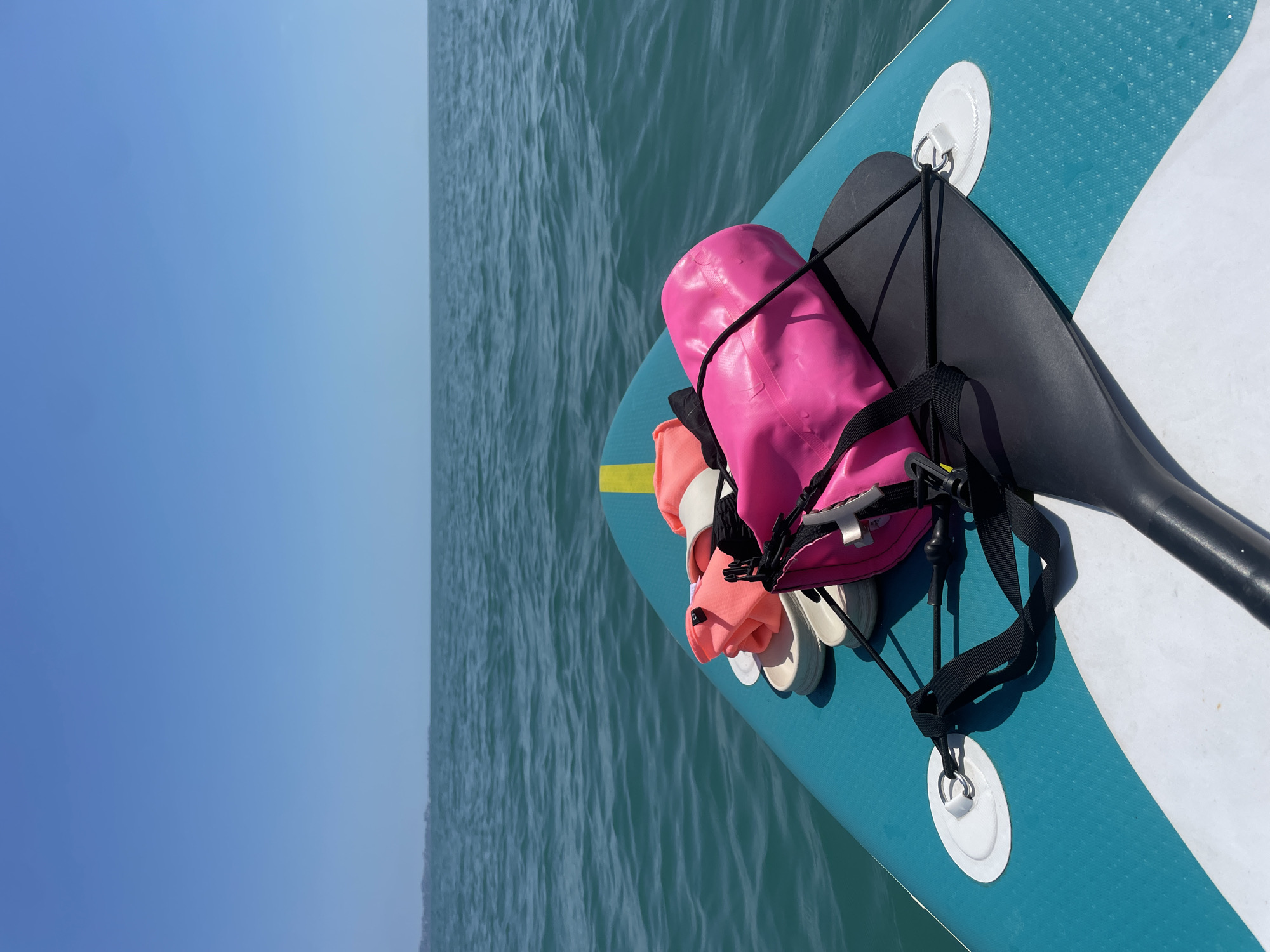 A first-person perspective shows the front of a turquoise and white stand-up paddleboard on a calm, blue ocean under a clear sky. On the paddleboard are a black paddle, a pink dry bag, a pair of white flip-flops, and an orange item of clothing or fabric. The horizon line is visible in the distance, separating the sea from the sky, with a faint coastline visible on the left. The scene appears to be located in Sochi, Russia, given the context.