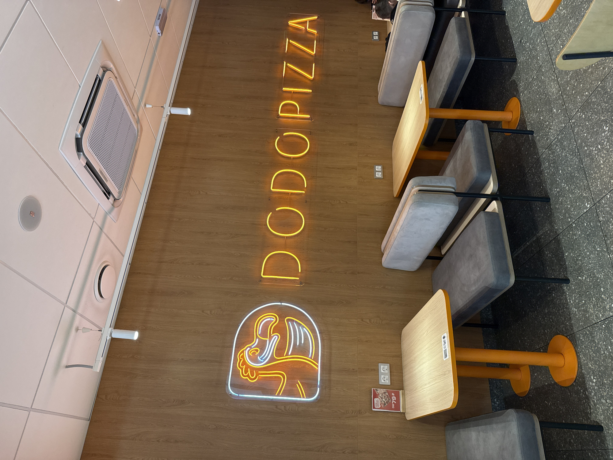 The image shows a top-down view of a Dodo Pizza restaurant. The scene includes wooden floor panels with the "DODO PIZZA" neon sign displayed vertically. A smaller neon logo is also visible. The seating arrangement consists of tables with orange supports and gray cushioned seating. The floor beyond the wooden section is tiled with a gray material. On the ceiling, there is an air conditioning unit and light fixtures. The image suggests a clean and modern fast-food restaurant interior. The location is likely in Tobolsk, Russia, given the Dodo Pizza brand association.