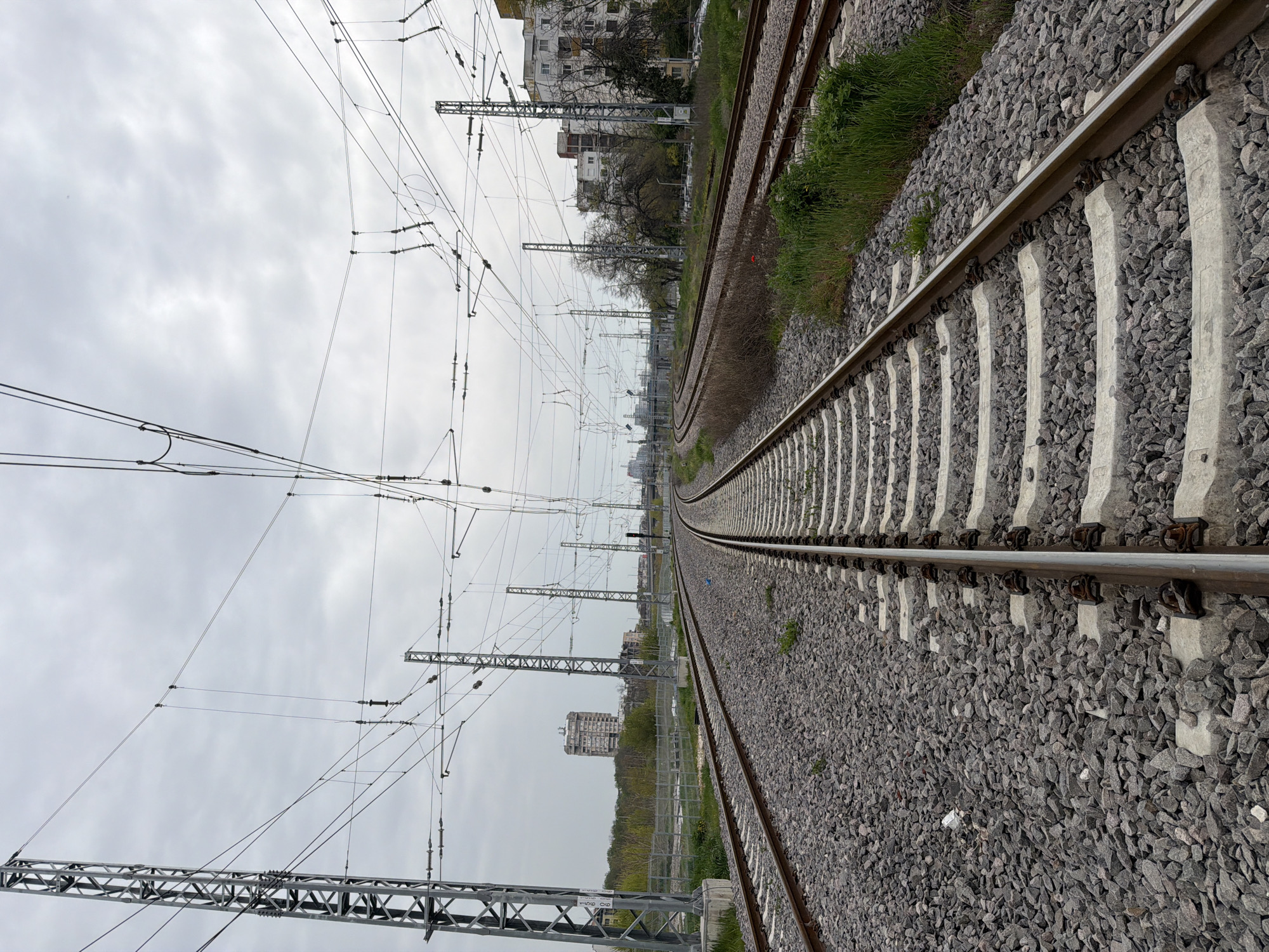 The image depicts a set of railroad tracks stretching into the distance under an overcast sky. The perspective is from ground level, looking along the tracks, which are made of steel rails supported by concrete sleepers. Small, dark grey ballast stones fill the spaces around the sleepers. Overhead, power lines and supporting structures are visible. In the background, buildings and trees appear, suggesting an urban or suburban location. Based on the prompt, this scene is located in the city of Plovdiv, Bulgaria. The image is static, and no people or actions are directly visible.