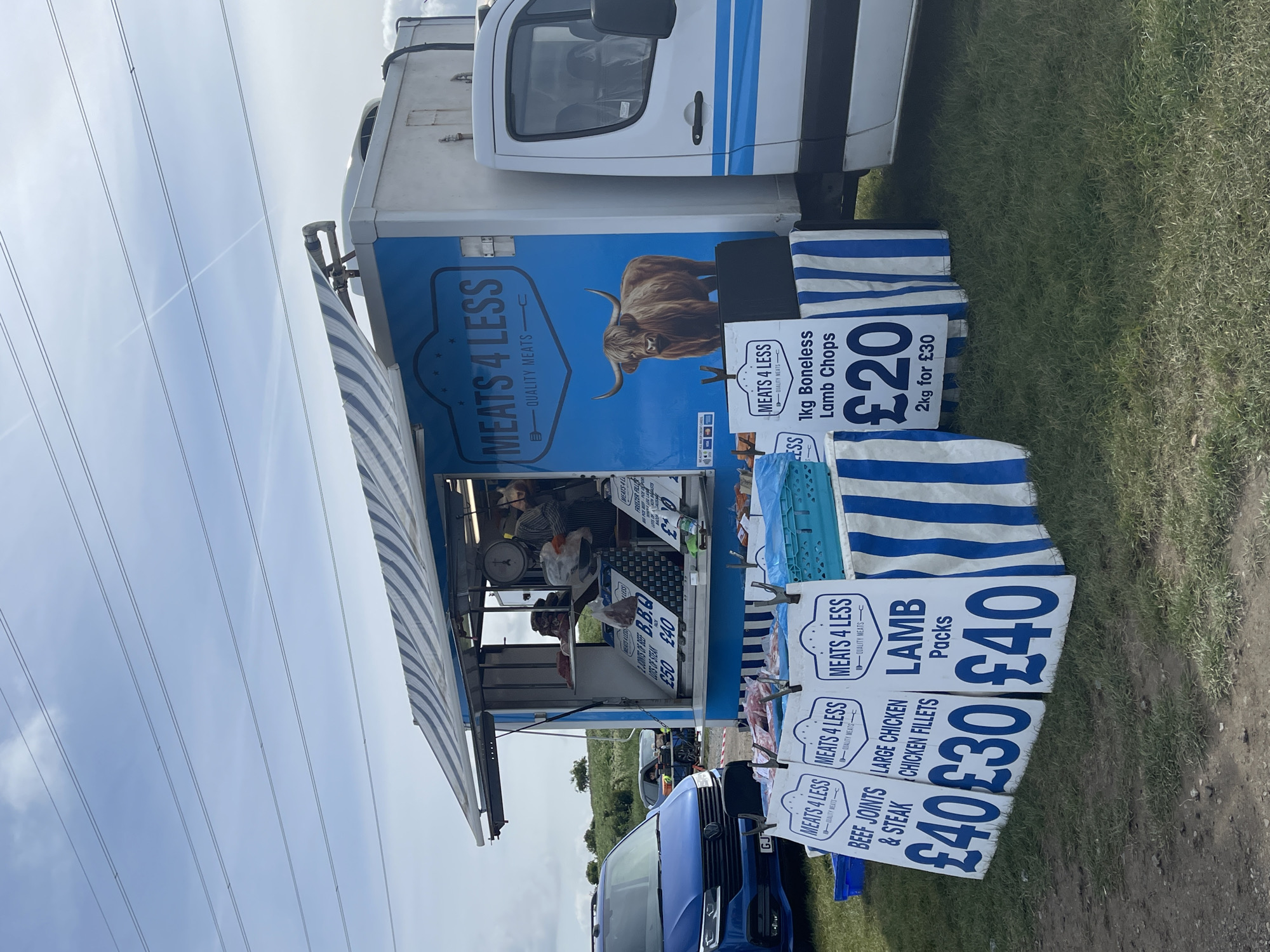 The image shows a "Meats 4 Less" food truck parked on a grassy area, likely a market or fairground. The location appears to be near Sevenoaks, United Kingdom, as indicated by the UK currency signs (£). The truck is white with a blue stripe and features a picture of a highland cow. Price signs advertising various meat products are displayed in front of the truck. The signs show prices for: 1kg boneless lamb chops at £20, 2kg for £30; Lamb packs for £40; Large chicken chicken fillets for £30; and Beef joints & steak for £40. A blue van is partially visible in the foreground. People are seen inside the truck. Overhead power lines are visible in the background, suggesting an outdoor location with nearby infrastructure.