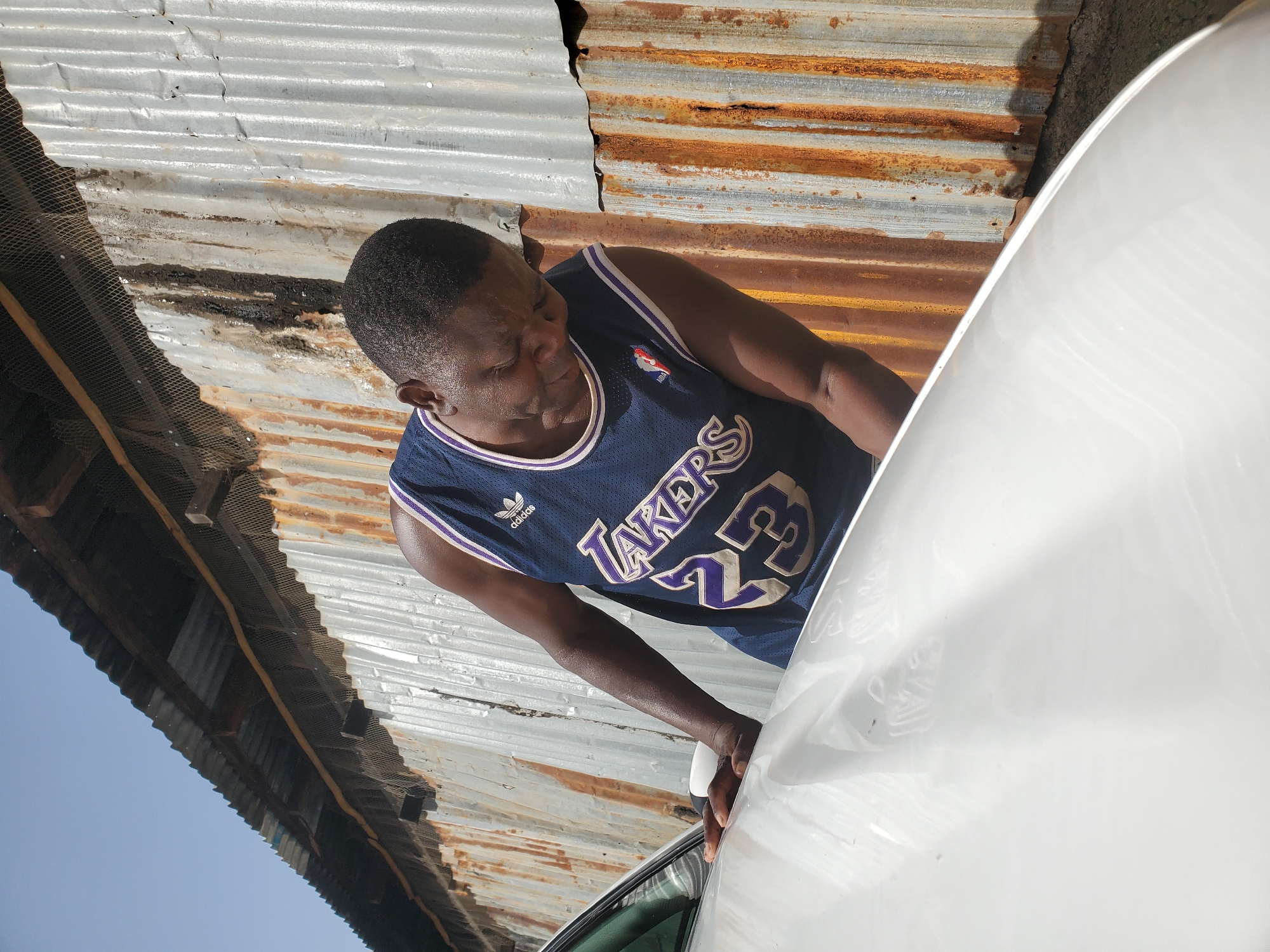 A man is leaning over the hood of a white car, with his head down. He is wearing a dark blue Lakers jersey with the number 23 on it. The background is a corrugated metal wall with rust and water stains. The sky is clear and blue. The scene appears to be in an outdoor urban environment, possibly in Bukuru, Nigeria. The lighting suggests it is daytime, with the sun casting shadows.