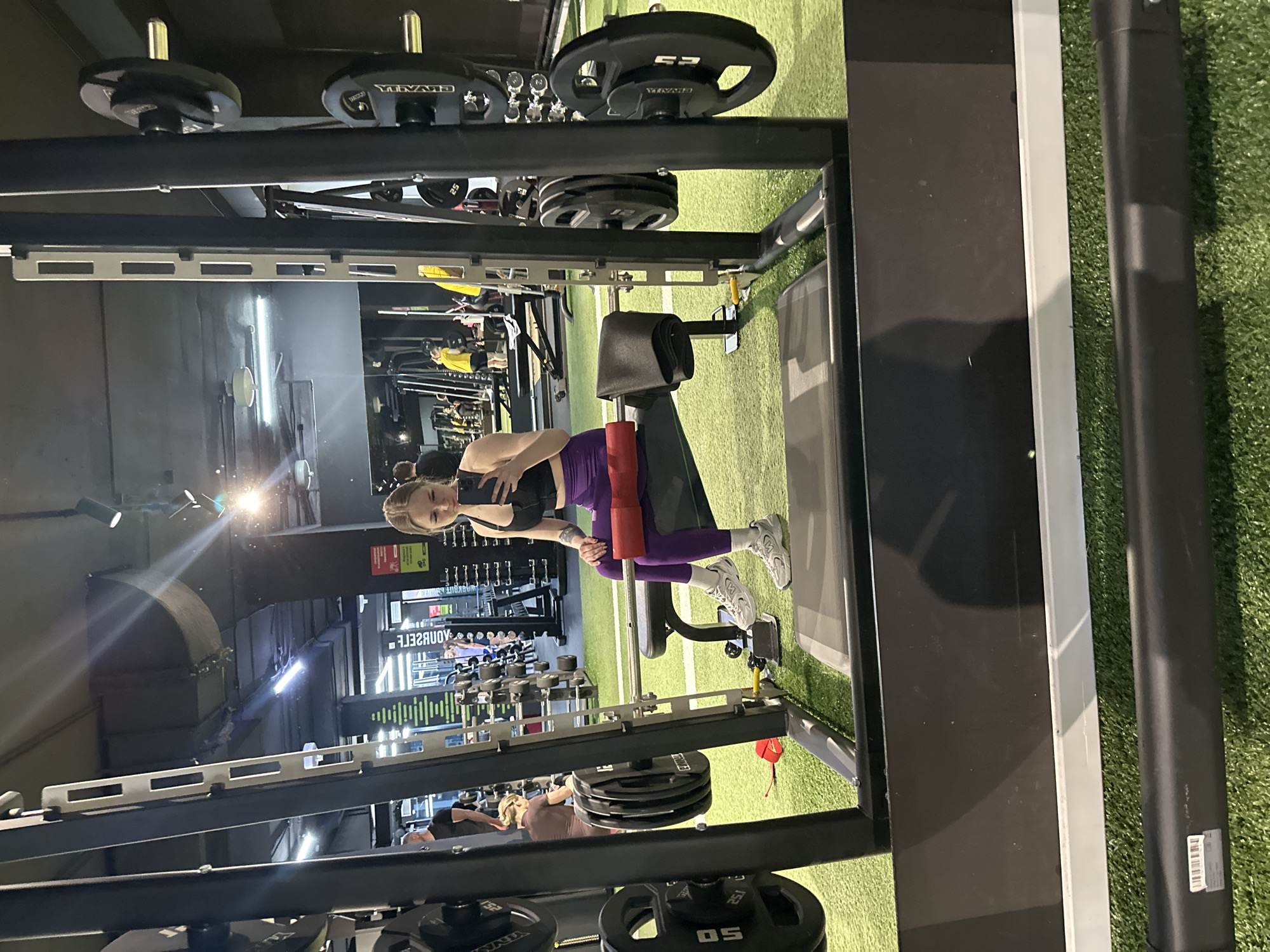 The image shows a young woman in a gym setting. She is wearing a black sports bra and purple leggings, and she is using a leg curl machine. The gym has artificial turf flooring and weight racks. The location appears to be an indoor gym, though the specific city and state are not discernible.
