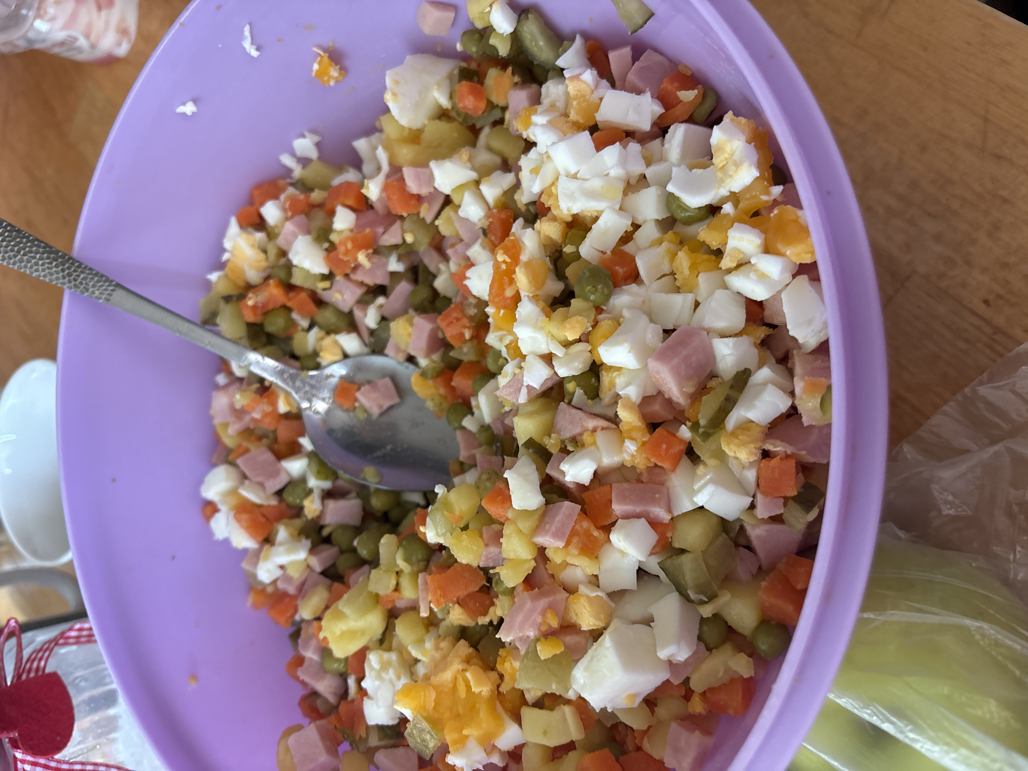 Here is a forensic description of the image:
A top-down shot depicts a large, purple plastic bowl filled with a mixed salad. The salad consists of diced ham, hard-boiled eggs, carrots, potatoes, pickles, and peas. A silver spoon rests on the salad within the bowl. The bowl is placed on a wooden surface. In the background, a white ceramic object and a decorative glass jar with a red and white ribbon are partially visible. The scene appears to be indoors.