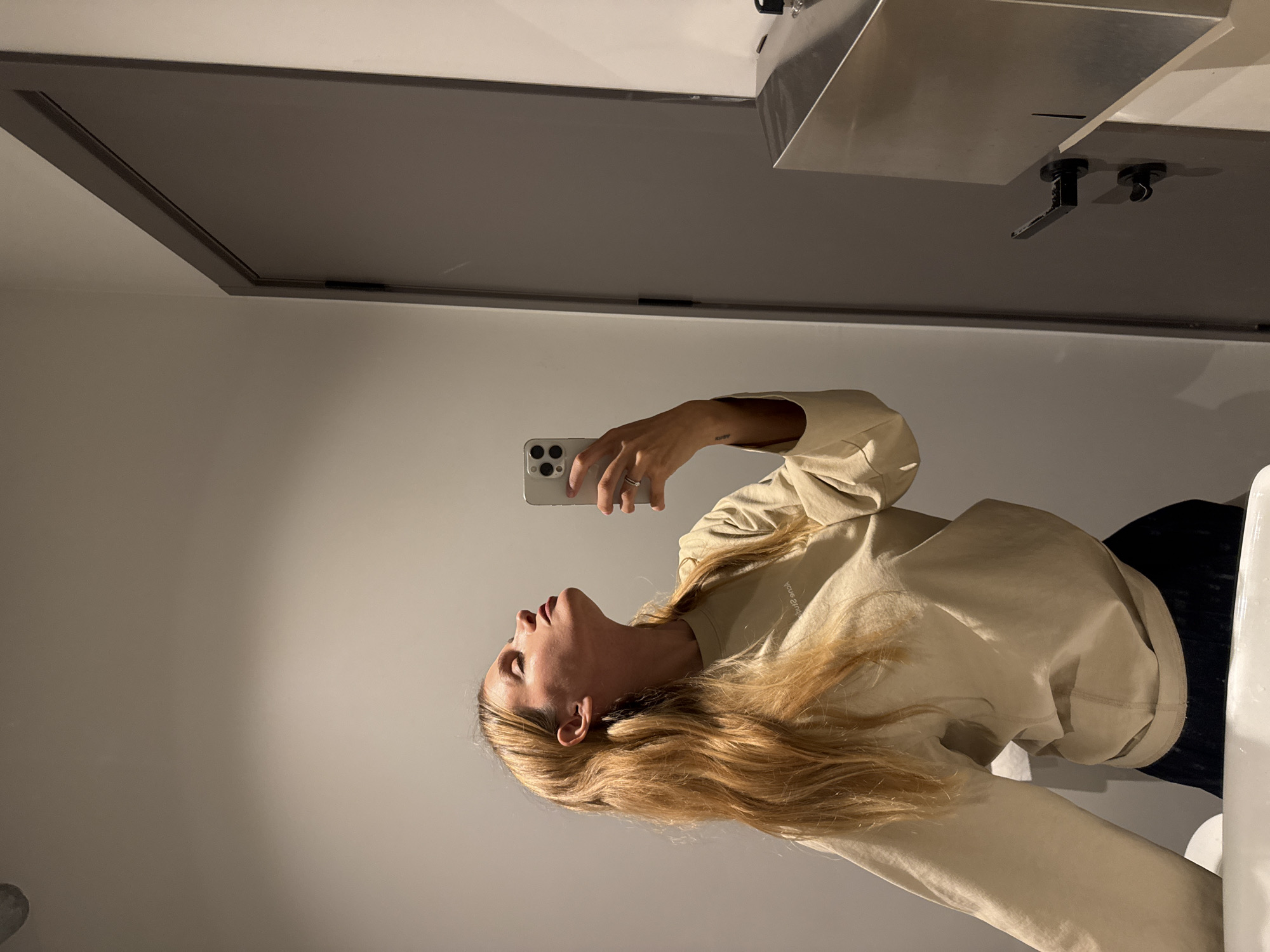 A young woman with long blonde hair and wearing a beige sweatshirt is taking a selfie in a bathroom. The bathroom has a grey door, light grey walls, and a stainless steel sink fixture. The location appears to be an indoor public restroom setting. There are no obvious identifying features to suggest a specific location in Tbilisi, Georgia.