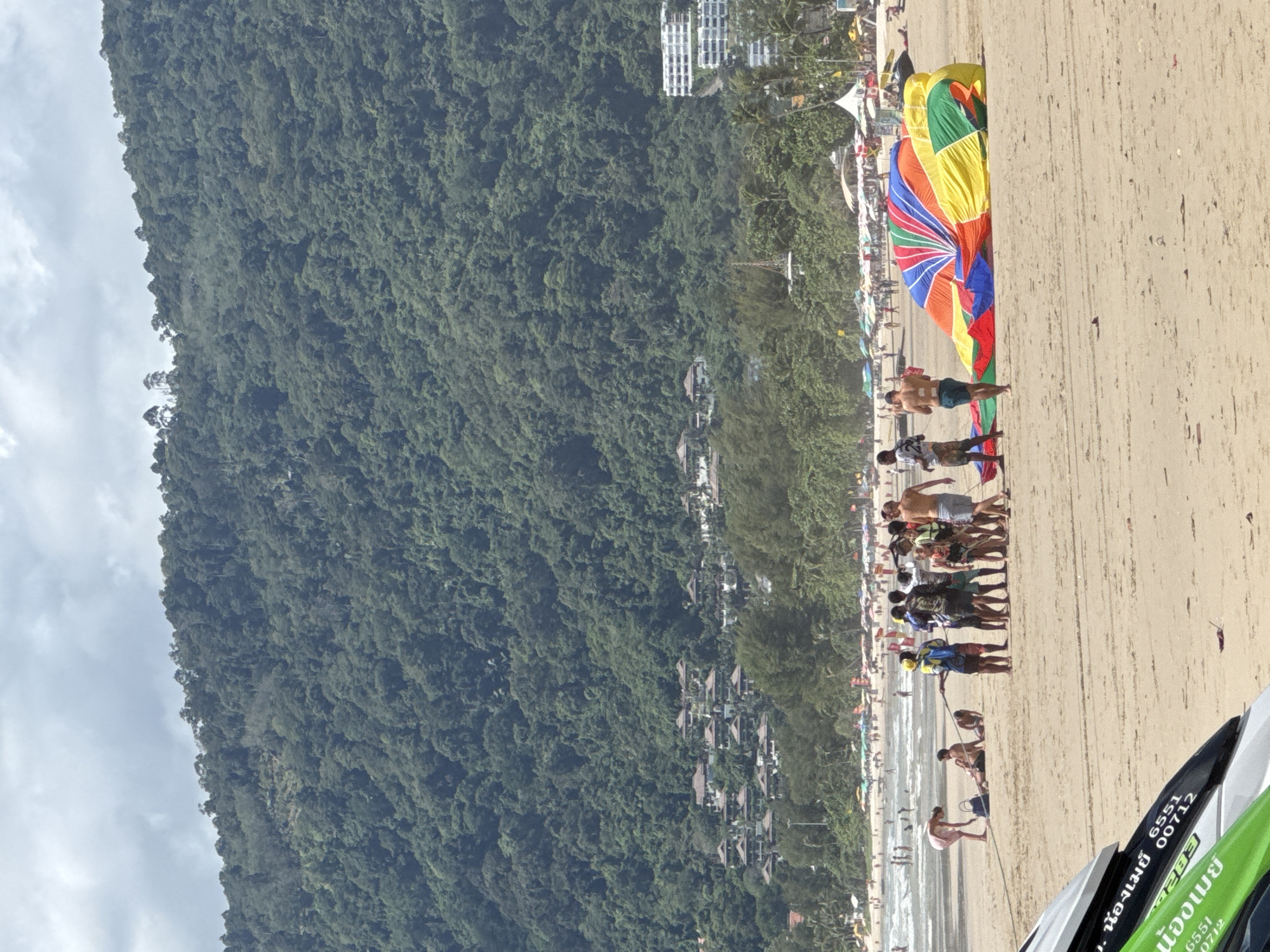 In Patong, Thailand, on a sunny day at the beach, a group of approximately 15 to 20 people are gathered around a deflated, brightly colored parachute on the sand. The parachute is predominantly red, blue, yellow, and green. Several individuals are standing, observing or possibly involved in packing up the parachute, while others are seated on the sand. In the background, the ocean is visible with a few waves, and a crowded beach with numerous umbrellas and sunbathers stretches along the coastline. Beyond the beach, a lush, green hillside covered in dense vegetation and dotted with buildings rises steeply. A few jet skis are partially visible in the foreground. The sky is cloudy.