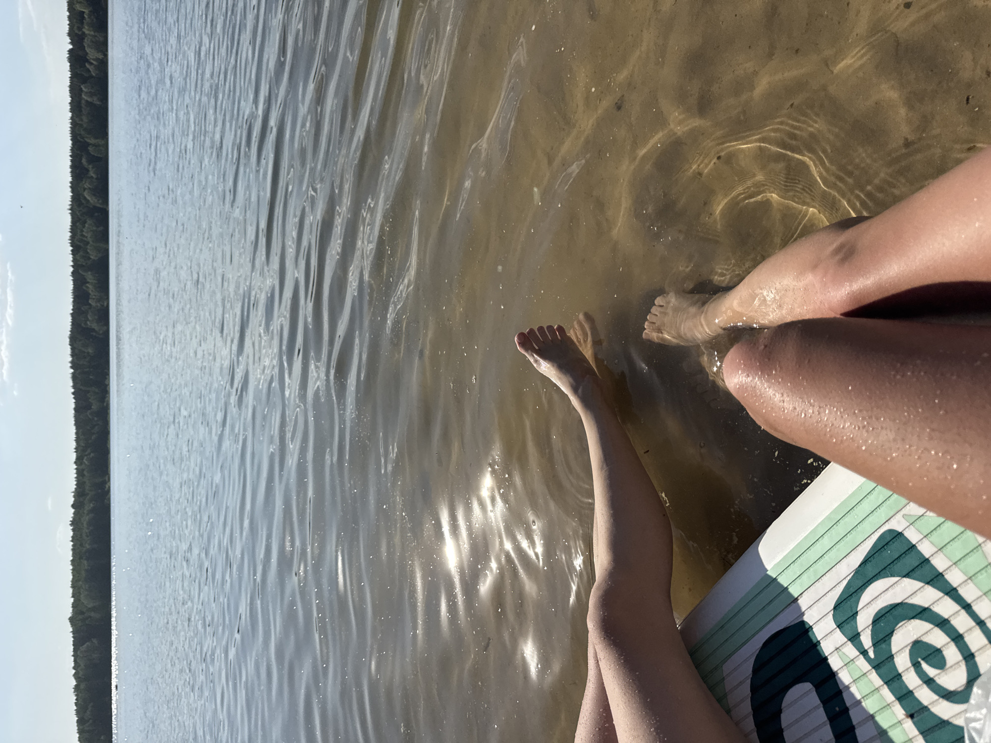The photograph depicts a view from the perspective of someone sitting on a paddleboard with their legs dangling in the water. The location is likely a body of water near Yal'chik, Russia, though this cannot be definitively confirmed. The water is relatively clear, and the sun's reflection creates shimmering patterns on the surface and the sandy bottom can be seen through it. Two pairs of legs are visible, partially submerged in the water, extending from the paddleboard. One paddleboard is partially shown with a blue and white swirly pattern design on it. In the background, a line of trees appears to be along the shoreline. The overall impression is one of a relaxed, sunny day at the water.
