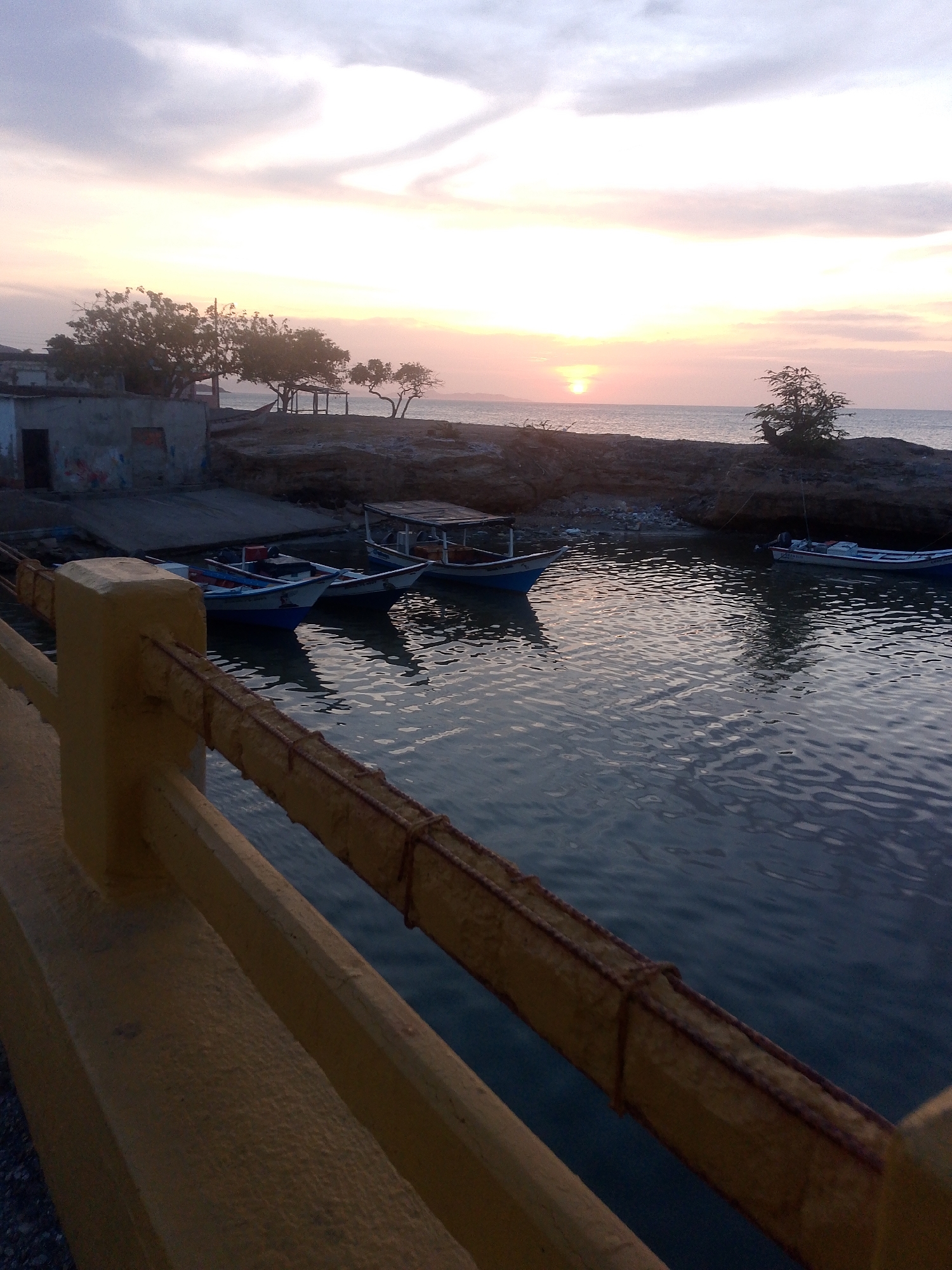 The image depicts a waterfront scene in Juan Griego, Venezuela. The sun is setting over the ocean, casting a warm glow on the horizon. In the foreground, a yellow-painted barrier lines the edge of a harbor or inlet. Several small boats are moored in the calm water, reflecting the colors of the sky. Buildings and sparse vegetation line the shore. A concrete ramp leads down to the water, suggesting a boat launching area.