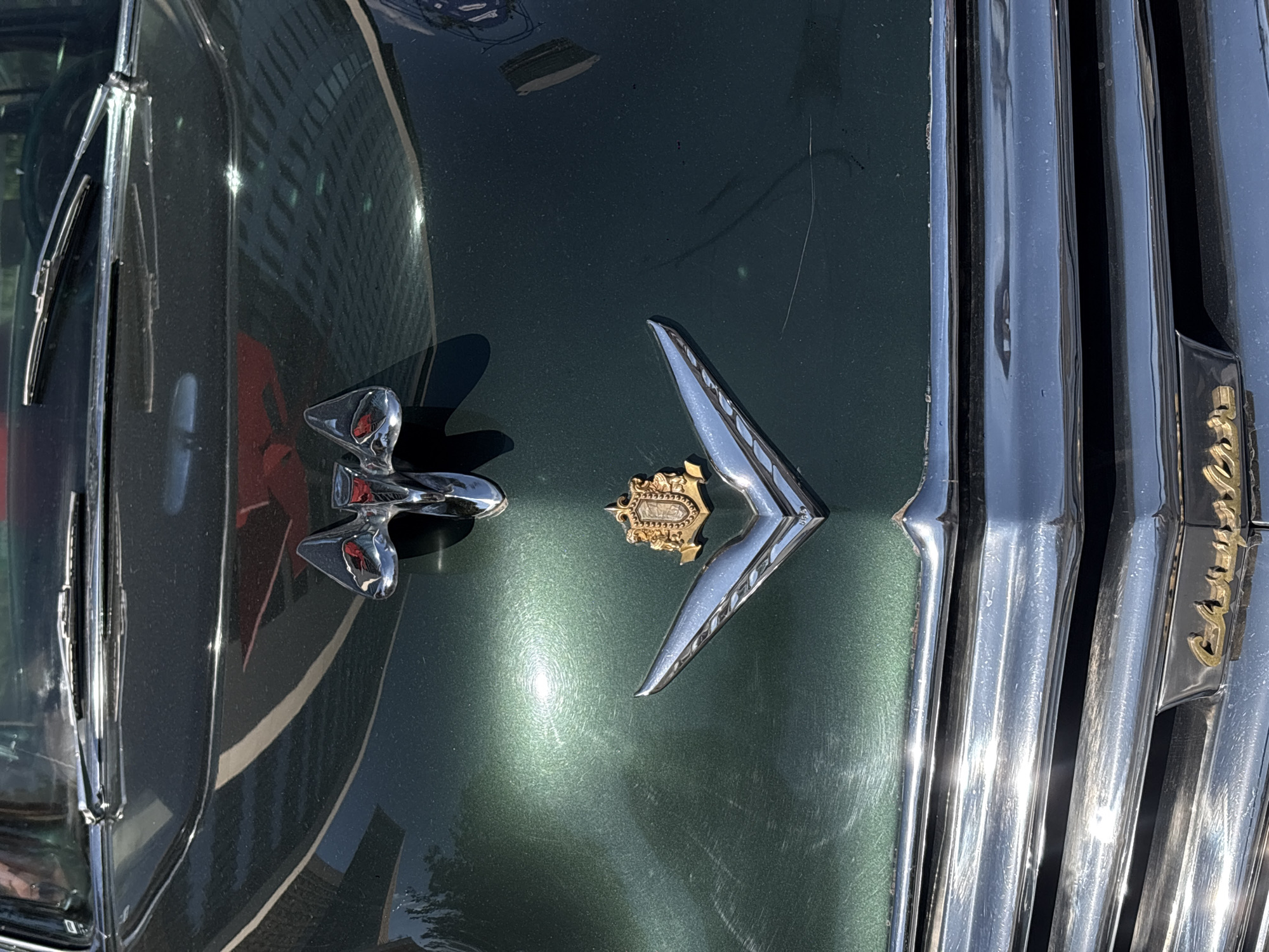 A close-up, slightly tilted shot shows the hood of a dark green vintage car. On the left side, a silver hood ornament resembling a stylized bird or plane is visible. Centrally, another silver and gold ornament, a winged "V" shape with a central crest, is positioned. To the right, part of the car's chrome grill is visible, with the word "Cadillac" in gold script on its right edge. The car's green paint has a glossy finish with some swirl marks. Reflections of buildings and a red object are visible on the paint. The image suggests the car is in an urban environment, likely in Mexico City. There are no people visible in the frame.