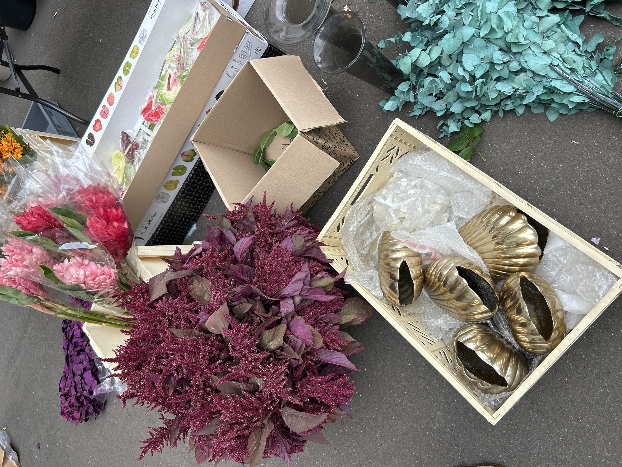 The image shows a collection of flowers and floral supplies scattered on an asphalt surface, likely outdoors. In the foreground, a cream-colored plastic crate contains five decorative gold objects, each with a ribbed, shell-like appearance and an open, hollow interior. These objects are nestled within bubble wrap. To the right of the crate, a large bunch of blue-green eucalyptus leaves is arranged. Behind this, a tall, dark glass vase stands upright. Further back, a cluster of deep red amaranth flowers with long, feathery plumes dominates the left side of the image. To the right of the amaranth, a white plastic crate holds several stems of bright pink and red ginger flowers, partially wrapped in clear plastic. In the background, a cardboard box contains a few green leaves and what appears to be a small package. Another opened cardboard box sits next to it. A partially visible cardboard box with printed images is stacked behind the ginger flowers. The ground is a dark grey asphalt. There are no people visible in the image. The presence of floral arrangements, crates, and boxes suggests a market stall, florist shop, or a delivery area. No specific landmarks in Berlin, Germany, are identifiable from the image.