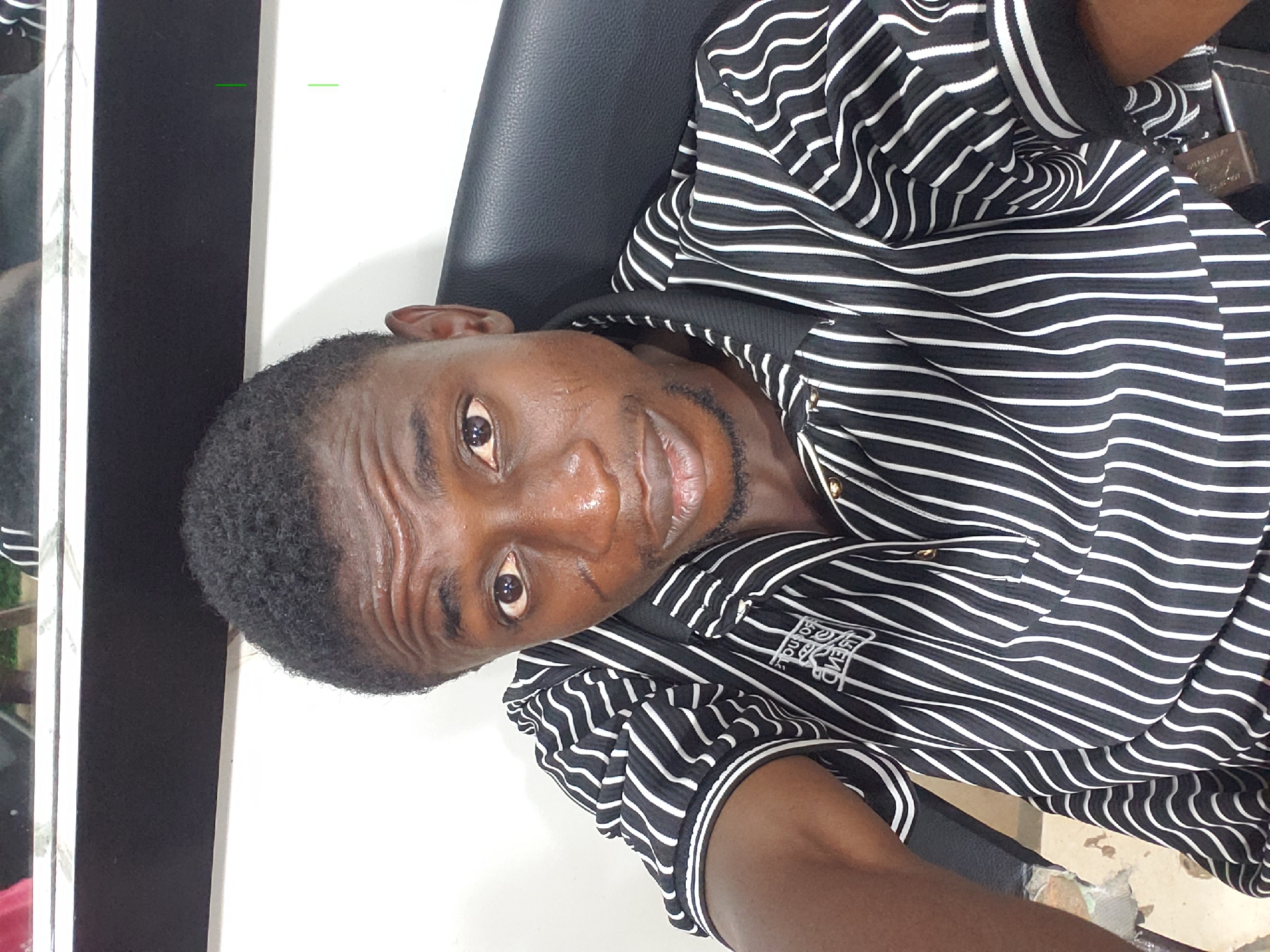 The media file captures a young man from the chest up, appearing to be of African descent, looking directly at the camera. He has short, dark, curly hair and a short, neatly trimmed beard. He wears a black and white horizontally-striped short-sleeved polo shirt with a black collar and white piping. Small gold-colored buttons are visible on the shirt's placket. On the right side of his chest, a white embroidered logo is present, with discernible text that includes "EST. 1978" and "CLUB".
The man is seated or leaning back on a black leather or synthetic chair. A brass-colored padlock, clearly inscribed with "SPECIAL ALLOY," is visible near his right shoulder, seemingly attached to a dark strap or part of the furniture. The background features a plain white wall on the right and a dark vertical structure, possibly a doorframe or partition, with a reflective edge on the left. Through the reflective surface, hints of greenery and what might be a vehicle are faintly visible. The man's expression is neutral to slightly smiling. The scene suggests an indoor or semi-enclosed setting, likely during the daytime. The location is Adankolo, Nigeria.