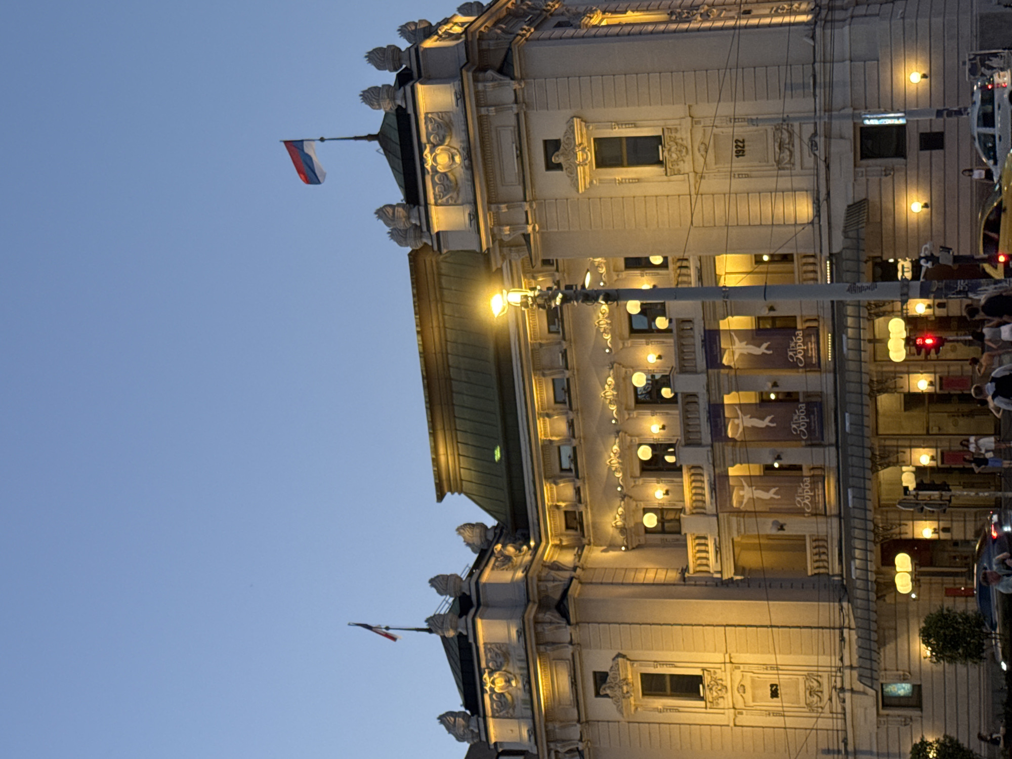 The photograph captures the National Theatre in Belgrade, Serbia, at dusk. The imposing neoclassical building is illuminated by warm, artificial lights, highlighting its ornate facade, including decorative sculptures, windows, and balconies. A Serbian flag flies prominently from the building's roof. In front of the theatre, a street is visible with a traffic light showing red, indicating a pause in traffic. Several cars are present on the street, and a few pedestrians can be seen walking on the sidewalk. The sky above is a clear, deep blue, characteristic of twilight. The overall scene conveys a sense of grandeur and activity in an urban setting during the evening.