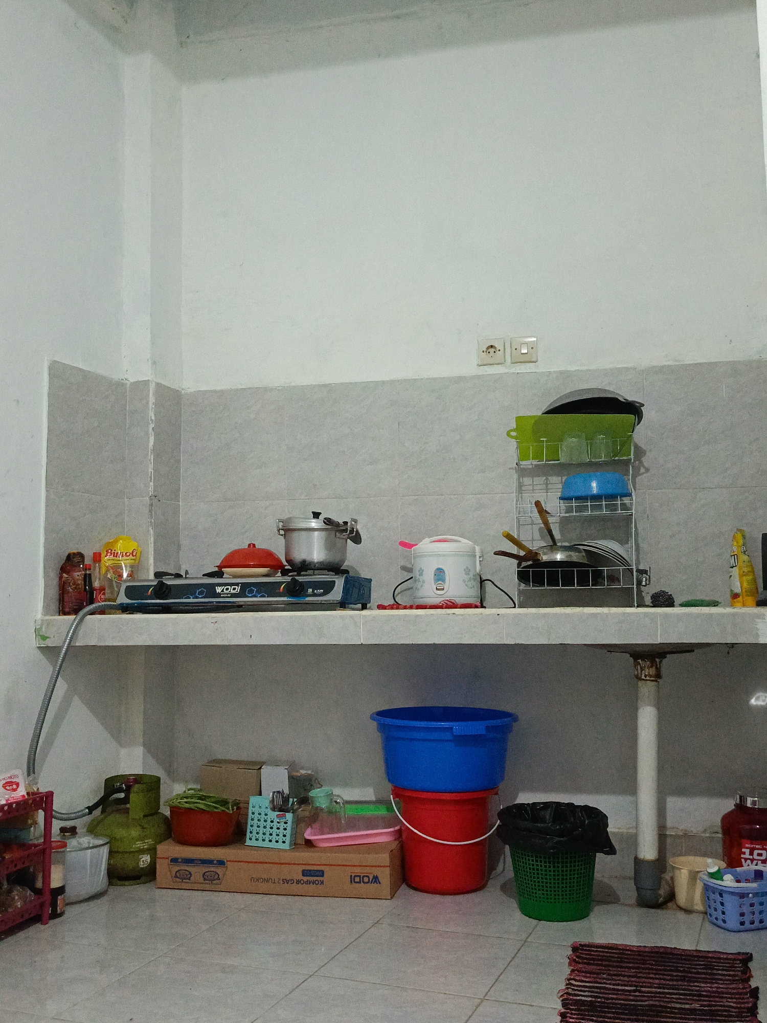 The image shows a modest kitchen in Bonder, Indonesia. The walls are tiled with a grey and white pattern, and the floor is tiled with light grey squares. On a white shelf, there is a gas stove with various cooking pots and pans. To the right, a dish rack holds more kitchenware, including bowls and glasses. A rice cooker is also visible on the shelf. On the floor, there are several buckets, a green gas cylinder, and storage boxes. A red bottle and a blue basket containing items are also on the floor. The overall impression is that of a functional, but somewhat cluttered, kitchen.