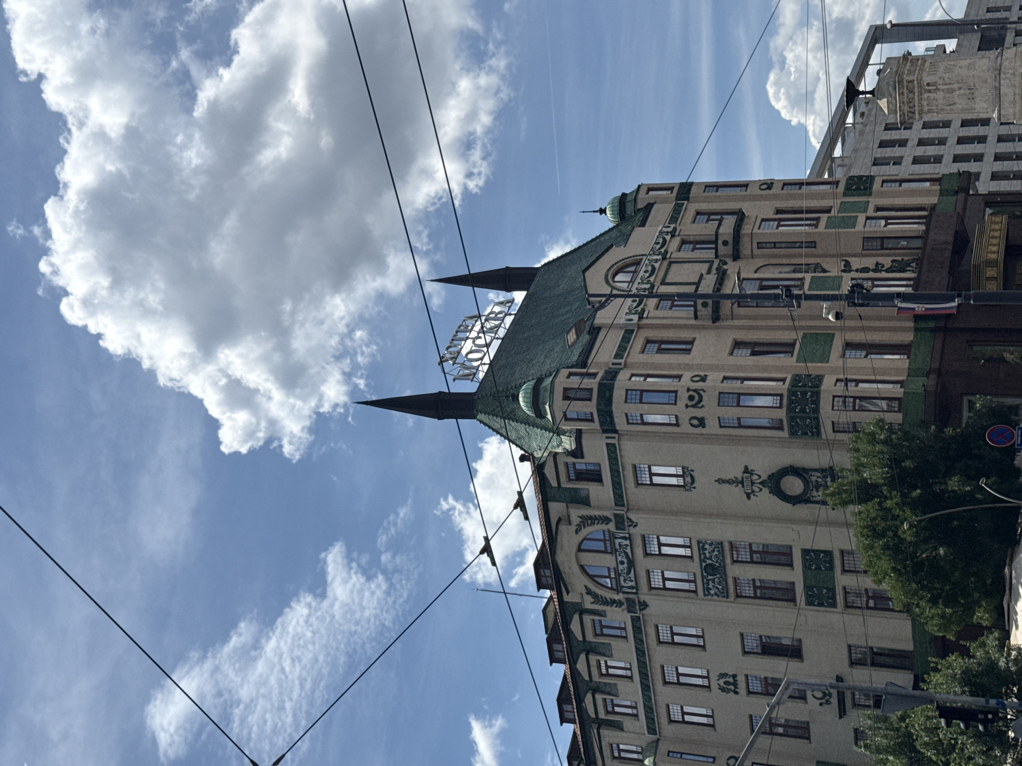 A historical, multi-story building with a green mansard roof and two spires is the main subject of this image, set against a bright blue sky with scattered white clouds. The building's facade is a light beige color, adorned with decorative green elements and numerous windows. A sign on the roof reads "HOTEL MOSKVA" in illuminated letters. Overhead, several black wires crisscross the frame, suggesting the presence of an urban transportation system, likely trams or trolleybuses, common in Belgrade. The architecture suggests an early 20th-century European style. To the left, a traffic light and trees are visible, indicating a street-level perspective. To the right, a modern building and a monument or sculpture with a column are partially visible, hinting at a location with a mix of old and new urban development. The overall impression is of a sunny day in a city with a rich architectural history. The building is Hotel Moskva in Belgrade, Serbia, located in the city center on Terazije Square.