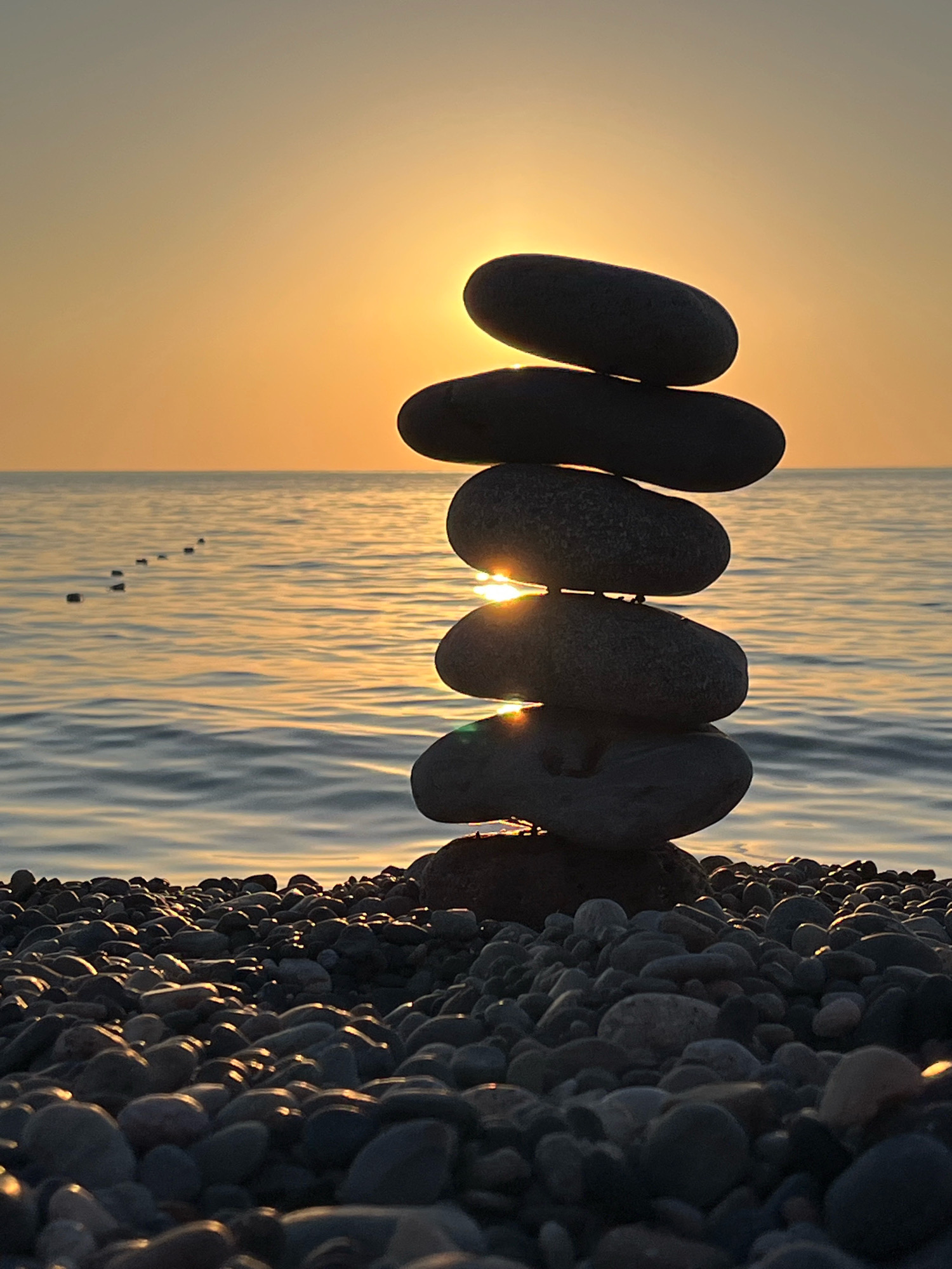 A stack of dark, smooth stones is balanced precariously on a pebble beach, silhouetted against a hazy orange sunset over the Black Sea. The sun is partially obscured by the top two stones, casting a warm glow and creating a lens flare effect. In the distance, a line of small, dark objects, possibly buoys or markers, stretches across the water. The beach is covered in an array of smooth, dark pebbles of various sizes. The scene is set in Kobuleti, Georgia.