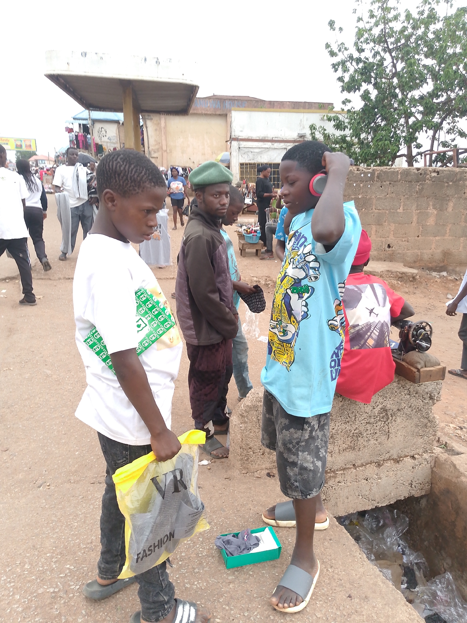 The image depicts a street scene in Bukuru, Nigeria. Two young boys are the central figures. The boy on the left is wearing a white t-shirt with a green patterned design, dark jeans, and sandals. He is holding a yellow plastic bag with the word "FASHION" printed on it. The boy on the right wears a light blue t-shirt with cartoon graphics, camouflage shorts, and sandals. He has red headphones on his head.
In the background, a man in a green beret, brown jacket, and maroon pants stands next to the boys. Another figure in red is seen seated on a concrete block behind the second boy, possibly operating a sewing machine. Other people are visible in the distance. The environment includes a concrete structure resembling a low wall, a gutter filled with plastic waste, and various buildings and objects typical of a street scene.