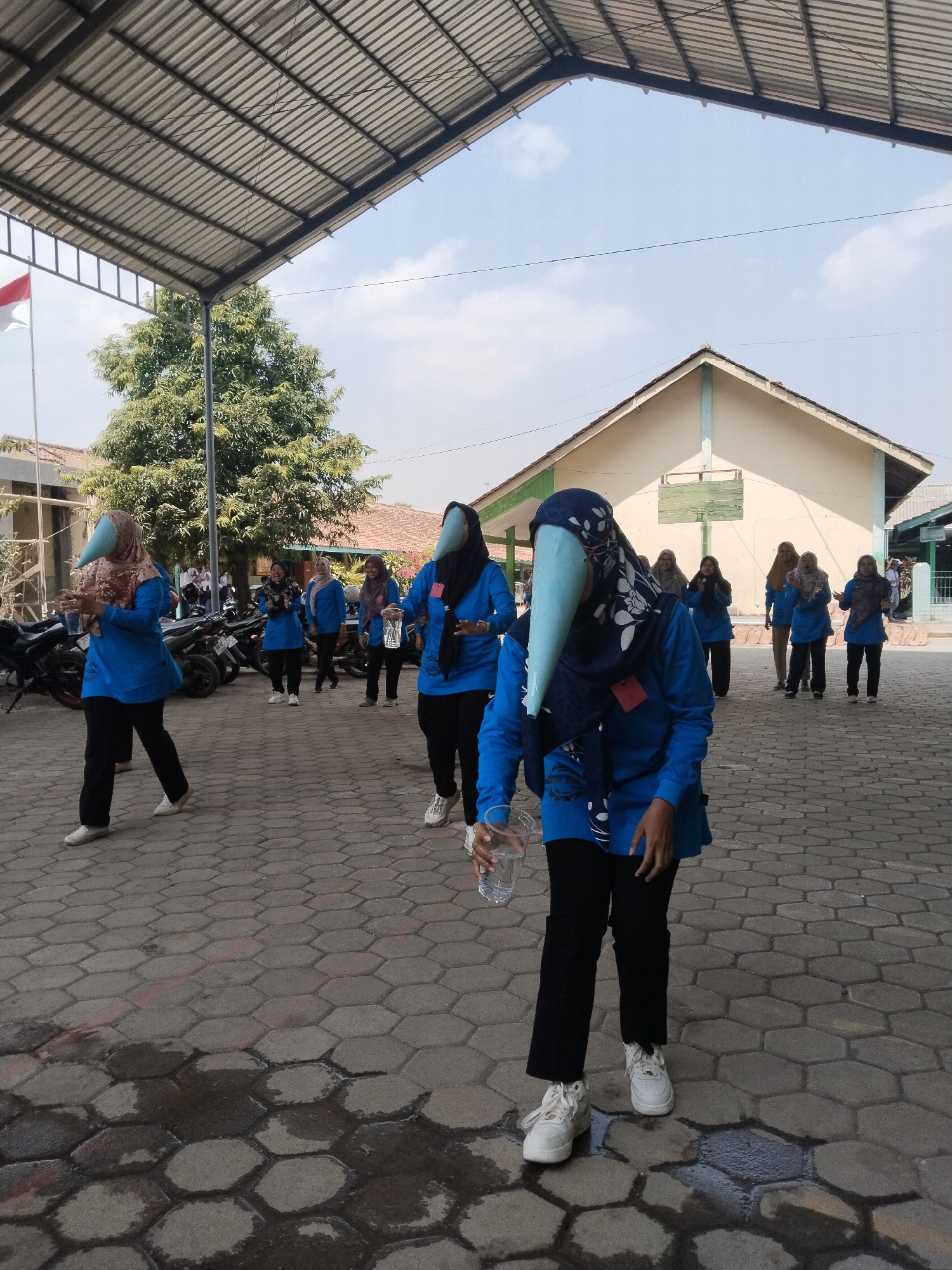 The image depicts a group of people, predominantly women, wearing blue shirts, black pants, and headscarves. Several individuals are wearing distinctive, light blue, conical masks that cover their faces, resembling bird beaks. The setting appears to be an outdoor area with a paved, hexagonal-patterned ground, possibly a schoolyard or public space. In the background, there is a building with a light-colored facade and a red-tiled roof, and a partially visible Indonesian flag on a flagpole. There are also motorcycles parked in the background. The sky is visible and appears cloudy. The overall scene suggests an outdoor event or activity. The provided location, "Pakulaut, Indonesia," is a coastal village in the Gresik Regency of East Java, Indonesia.