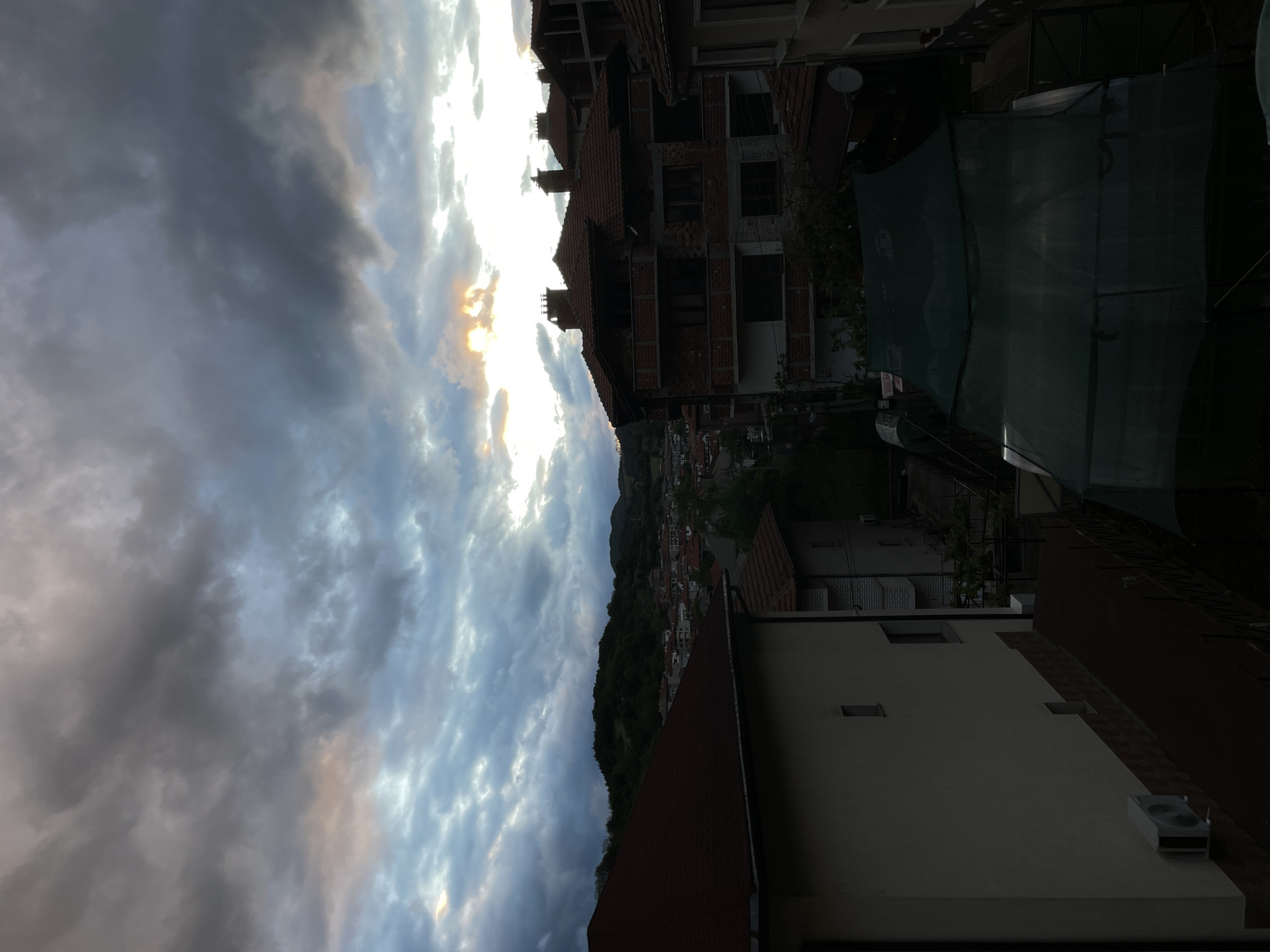 The image shows a view of Zlatograd, Bulgaria from an elevated position, possibly a rooftop. The sky is filled with dramatic clouds, with some sunlight breaking through. A few buildings are visible in the foreground, including a partially constructed brick building with a red tile roof and a white building with an air conditioning unit on the roof. A hillside with green vegetation and houses is located in the middle ground, blending into the town's rooftops. A tall green fence is also visible near the building. The overall scene depicts a blend of urban and natural landscapes under a dynamic sky.
