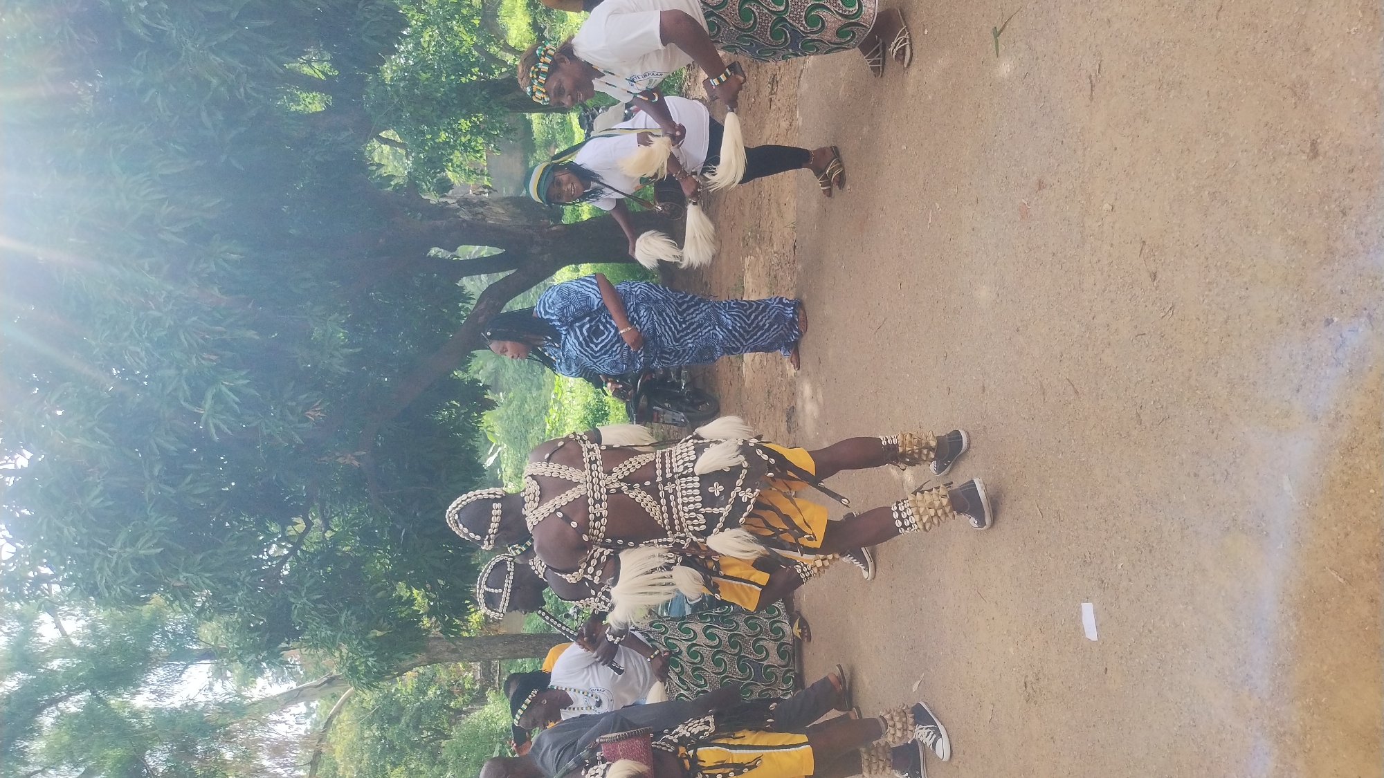 In Jos, Nigeria, this image captures a group of people in an outdoor setting, likely a street or a clearing, under the dappled sunlight filtering through trees. The central figures are two men in elaborate traditional attire, featuring ornate headdresses, shell necklaces, and skirts adorned with white tassels and shell embellishments. They are also wearing ankle decorations and dark sneakers. Their backs are to the camera, suggesting they are engaged in a performance or procession.
To their right, two women in white tops and patterned skirts are holding and fanning white plumes, also appearing to be part of the event. One of these women is wearing a green and yellow headwrap. Further to the right, another woman stands in a blue and white patterned dress. In the background, a motorcycle is parked, and more trees and foliage suggest a semi-rural or park-like environment within the city. The ground is a dusty, light-colored surface. The overall atmosphere seems festive and celebratory.
