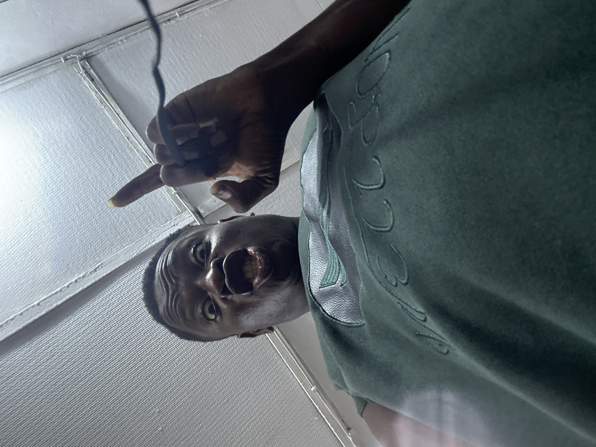 An individual with dark skin, likely male, is captured in a dynamic low-angle, tilted photograph. The subject's face is prominent, looking directly into the camera with a wide-open mouth, suggesting a shout, gasp, or intense expression, and wide eyes. Their right hand is raised, with the index finger extended and pointing towards the upper left side of the frame. The index fingernail has a light-colored polish.
The person is wearing a dark green long-sleeved shirt or hoodie, featuring stylized embroidered or embossed text on the front, parts of which resemble "VEN" and the digits "22". A thin, dark cable or wire hangs vertically in the background, near the raised hand. The setting appears to be an indoor space, with a light-colored, textured ceiling or wall visible behind the subject. The background features distinct seams or panels, indicating a structured overhead surface.
The image, taken in Ikorodu, Nigeria, conveys a moment of strong emotion or exclamation from the subject. The extreme low-angle perspective adds to the dramatic and immediate feel of the shot.
