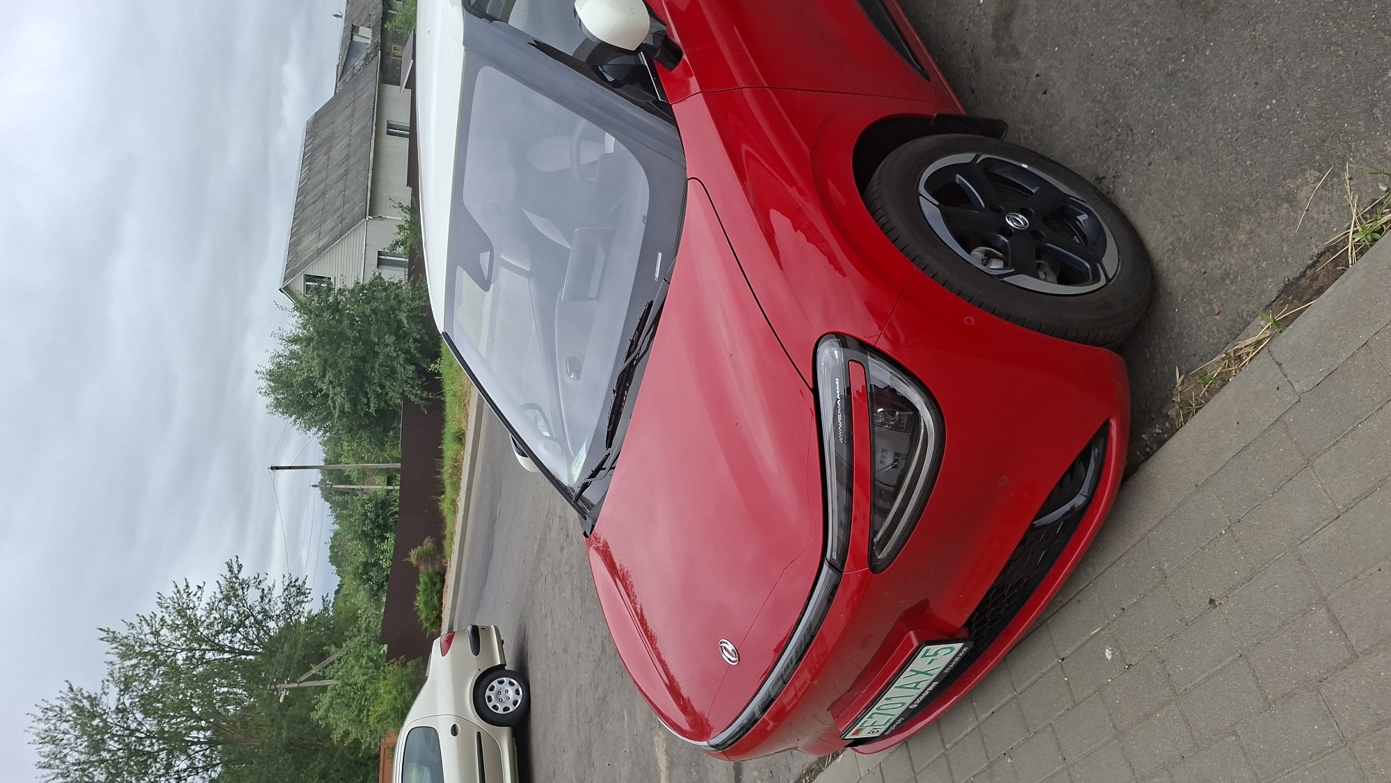 A bright red electric car with a white roof is parked on a sidewalk next to a street. The car has a license plate that reads "BY E701AX-5". In the background, there is a light-colored car, trees, houses, and a fence. The sky is overcast. The scene appears to be in a residential area of the city of Smalyavichy, Belarus.