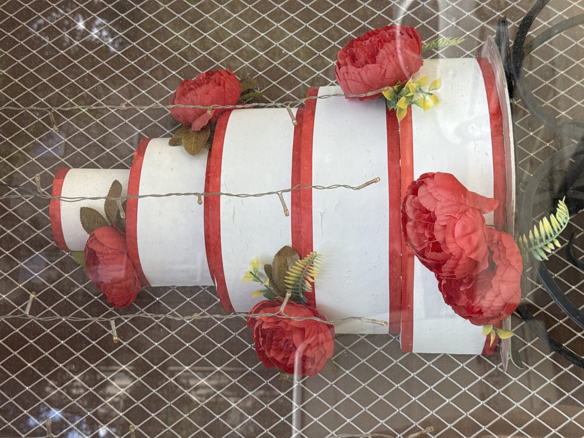 A five-tier, faux cake with white frosting and red ribbon accents sits behind a diamond-patterned wire mesh. Red artificial flowers and greenery are attached to the cake. String lights with clear bulbs are draped around the cake and mesh. The cake is displayed in a window, with a dark, mottled background visible through the mesh and cake.