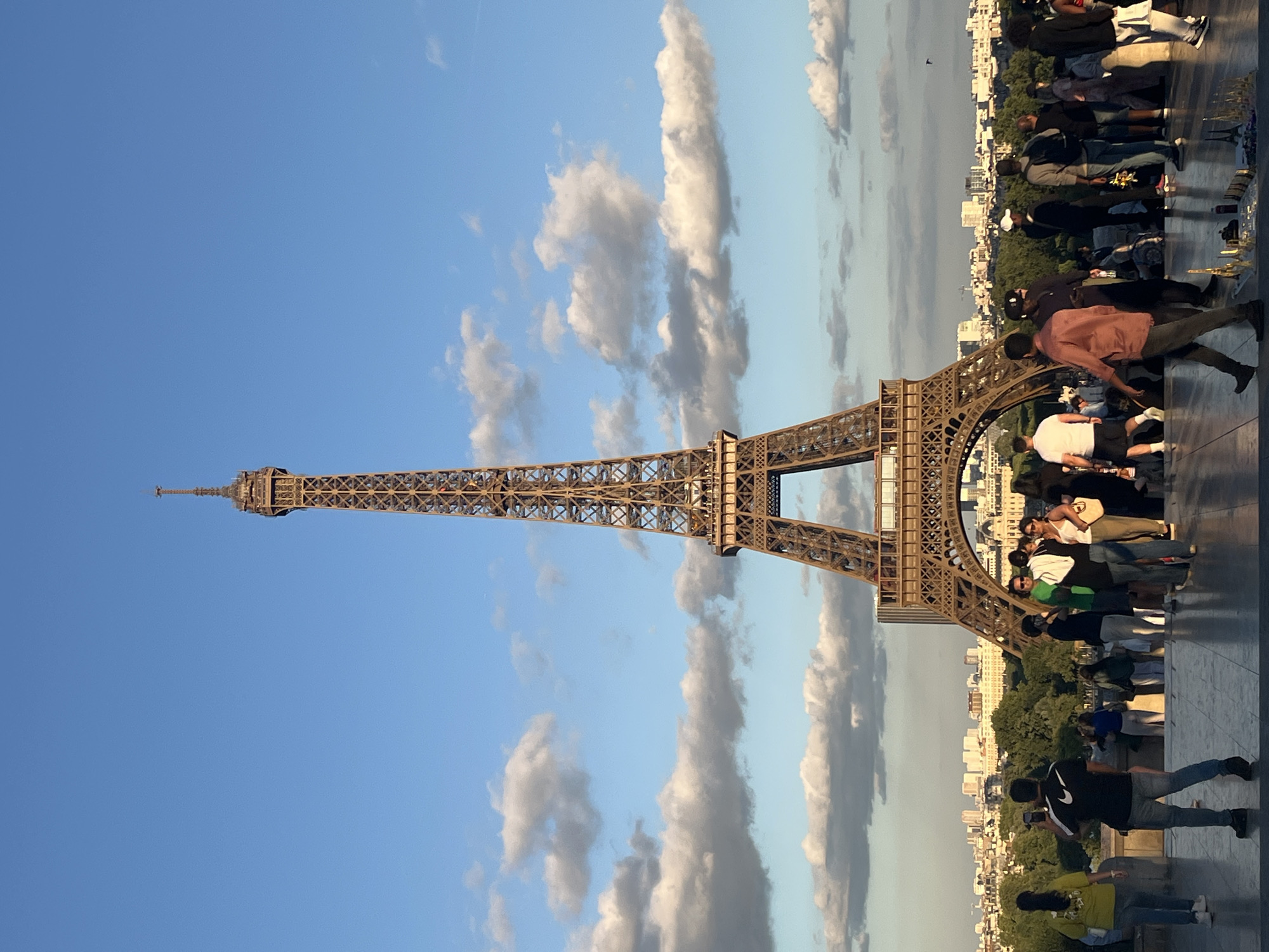 In Paris, France, the Eiffel Tower is prominently displayed against a backdrop of a bright blue sky with scattered white clouds. The tower, a latticework of brown iron, dominates the center of the frame. In the foreground, a group of tourists is gathered on a paved area. The people are engaged in various activities: some are looking at the tower, others are taking pictures, and a few are standing and conversing. The individuals are dressed in casual clothing. Behind the tourists, a panorama of Paris is visible, featuring a dense collection of buildings with trees interspersed throughout the urban landscape. The lighting suggests it is either early morning or late afternoon, with the sun casting a warm glow on the scene.