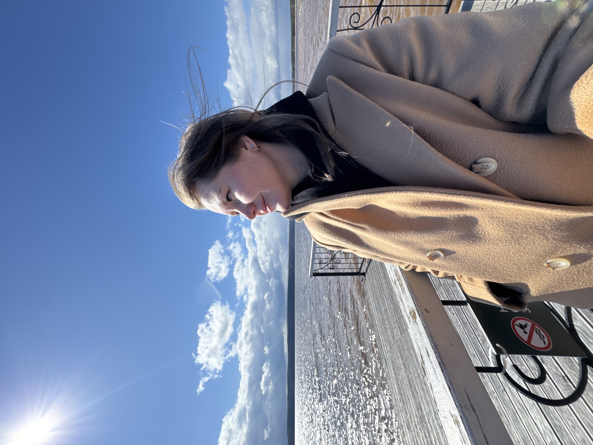 This vertical image features a young woman standing outdoors on what appears to be a pier or walkway alongside a large body of water. She is positioned to the left of the frame, looking gently downwards with a serene and contemplative expression. Her medium brown hair is subtly windblown, and she wears a light beige or tan coat over a dark garment, with a small silver earring visible in her left ear.
Behind and to the right of the woman, a vast expanse of water stretches towards a distant horizon, sparkling intensely under bright sunlight. The reflections on the water indicate a sunny day. A wooden pier runs along the right side of the image, featuring decorative black wrought-iron railings.
The upper and left portions of the image are dominated by a clear, bright blue sky dotted with scattered, fluffy white clouds. The sun is very prominent in the lower-left corner, emitting strong rays and lens flare, confirming it's daytime.
On the pier, near the bottom right, a dark green sign with white text and two red prohibitory symbols is partially visible. One symbol depicts a stylized person with a red circle and a diagonal line through it, typically indicating "no entry" or "no swimming." Another symbol shows a wavy line crossed out, possibly signifying "no fishing" or "no wading."
The setting suggests a peaceful, sunny day by a large lake or river, consistent with the provided location context of Polazna, Russia. The bright weather, the calm demeanor of the subject, and the shimmering water create a tranquil atmosphere.