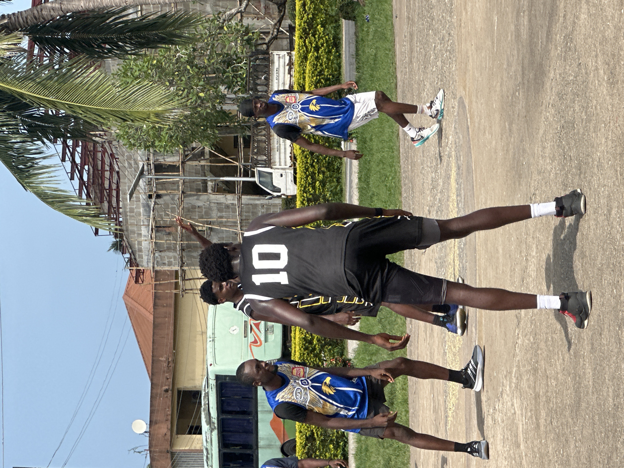 In Ajara, Nigeria, four young men are playing basketball on an outdoor court. The court appears to be paved with rough, light-colored concrete. In the background, a light green bus is parked. Beyond the bus, a building is under construction with visible scaffolding. A lush green hedge runs along the perimeter of the court, separating it from a grassy area. Two of the men are wearing blue and yellow basketball jerseys with eagles and "AFL" logos, while the other two are in black jerseys. One of the men in black has "10" prominently displayed on his jersey. The sky is clear and blue, suggesting a sunny day.
