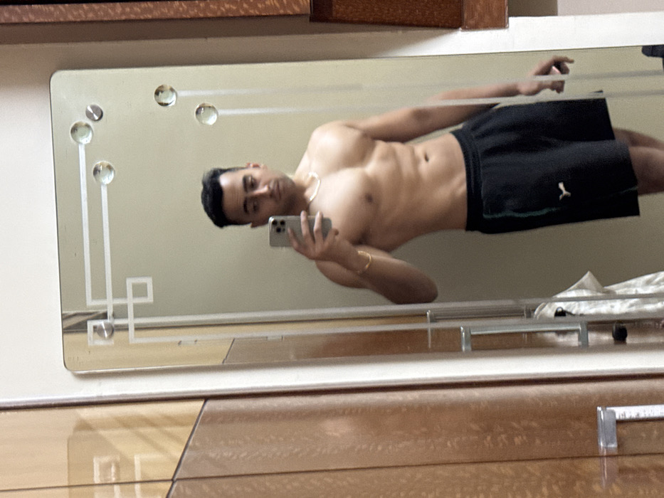 A young man, likely in his 20s or 30s, is captured in a mirror selfie taken indoors, possibly in a bedroom or dressing area in Kolkata, India. He is shirtless, displaying a lean and muscular physique with visible abdominal muscles and pectorals. He wears black athletic shorts with a white PUMA logo on the left leg, a silver chain around his neck, and a gold-colored bracelet on his left wrist. He holds a smartphone in his right hand, using it to take the photograph of his reflection. His expression is neutral, and he is looking directly towards the phone's camera.
The rectangular mirror in which he is reflected features decorative etched designs, including geometric lines and circular accents, particularly prominent on the top and bottom left sections. The background visible in the mirror is a plain, light-colored wall. Below the mirror, a piece of polished wooden furniture, possibly a dresser or wardrobe, is visible. It has a warm brown tone with a noticeable wood grain and features metallic fixtures. A white cloth or similar item is partially visible on this furniture to the right. The image captures a personal moment, showcasing the man's physique and casual indoor attire.