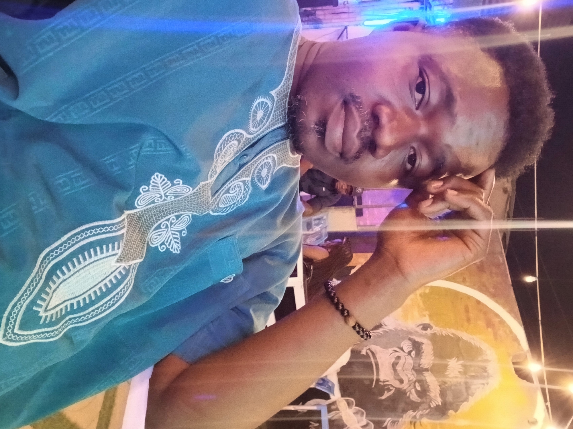 A man with a dark complexion, mustache, and short, curly hair is posing for a selfie. He is wearing a blue shirt with white embroidery around the collar. The background features a mural depicting a gorilla and a few other people seated. The location is identified as Jos, Nigeria. He is wearing a black bracelet with at least one tan bead. The photograph appears to have been taken outdoors, possibly at night, indicated by the overhead string lights and general lighting.