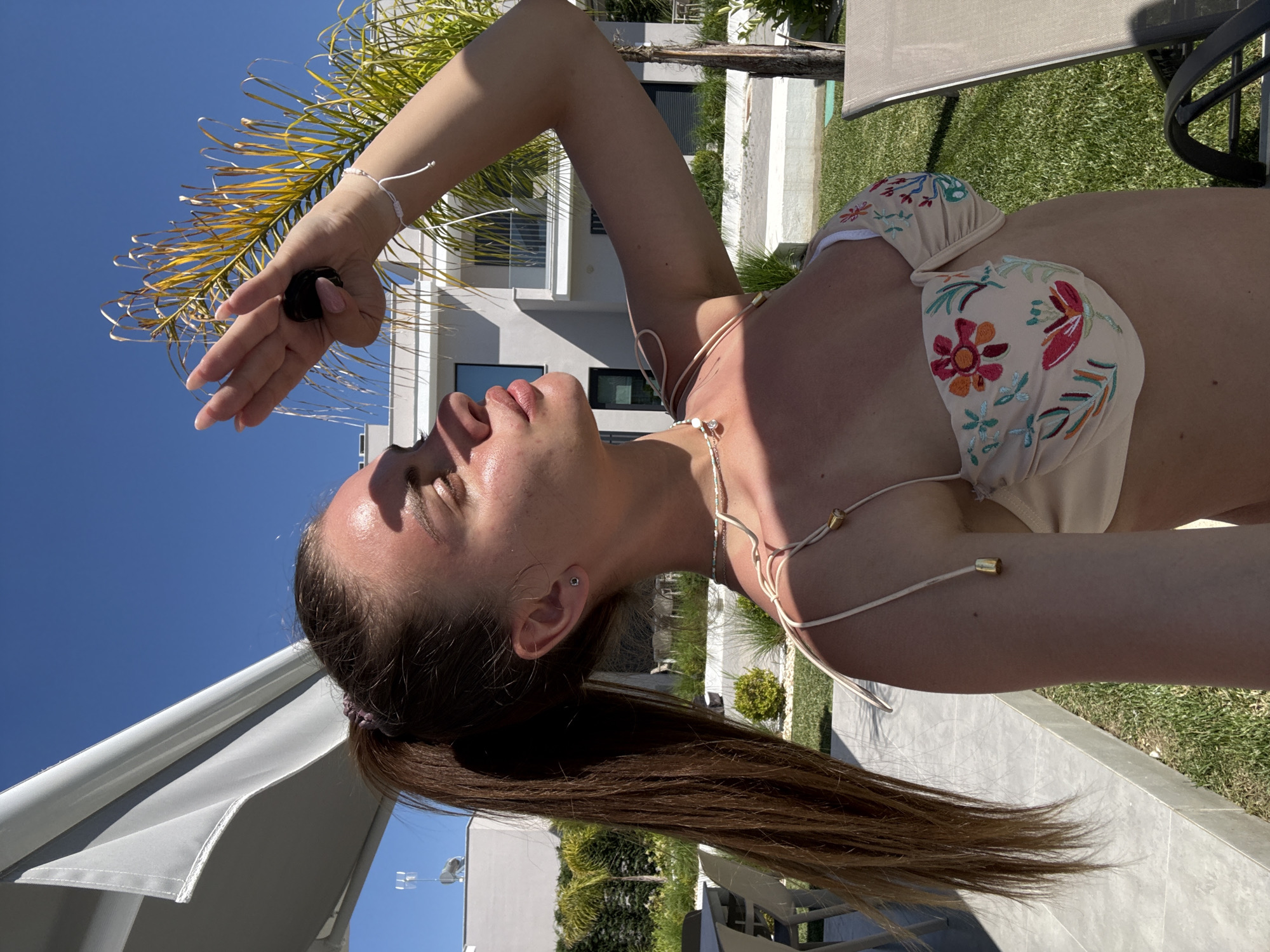A woman is lying on a chair outdoors under a bright sky, possibly in a location with palm trees, houses, and well-maintained lawns. She is wearing a light-colored bikini with floral patterns and a thin necklace. She is holding something in her hand, blocking the sun from her eyes. Her hair is pulled back in a ponytail. The location is Unknown, Unknown.