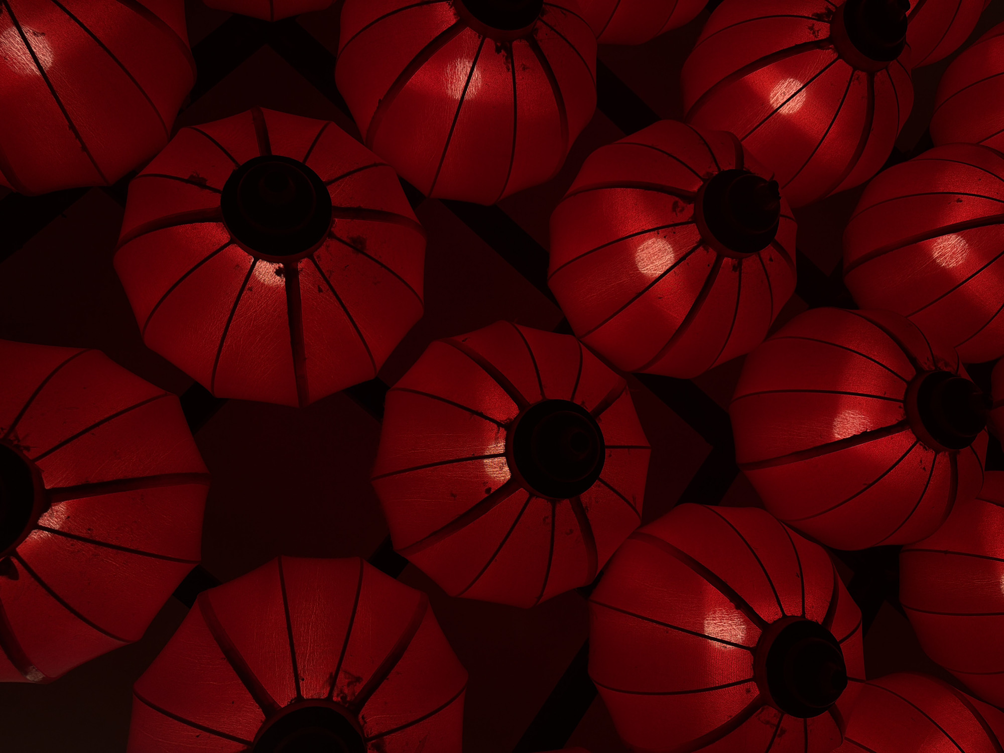 The image captures an overhead view of a dense arrangement of numerous red lanterns. Each lantern is spherical or squashed-spherical, constructed from a semi-translucent red fabric or paper stretched over an internal skeletal framework, creating visible segments. At the center of what appears to be the bottom of each lantern is a dark, circular cap or opening, likely where the light source or hanging mechanism is situated.
The lanterns are illuminated from within, emitting a vibrant, warm red glow that accentuates their texture and segmented structure. The internal light sources are visible as bright, circular spots on the surface of many lanterns. They are packed closely together, filling the frame with a rich pattern of red light and shadowed contours.
No people or specific activities are visible, but the illuminated display suggests a festive or decorative purpose. Given the location context of Đà Nẵng, Vietnam, these lanterns are characteristic of traditional Asian decor, frequently displayed during cultural festivals like Tết (Lunar New Year) or to adorn public and private spaces. The intense red color commonly symbolizes good fortune, prosperity, and happiness. The illumination indicates the scene is set during nighttime or in an interior space where the lanterns serve as the primary lighting and decoration. There is no visible text in the image.