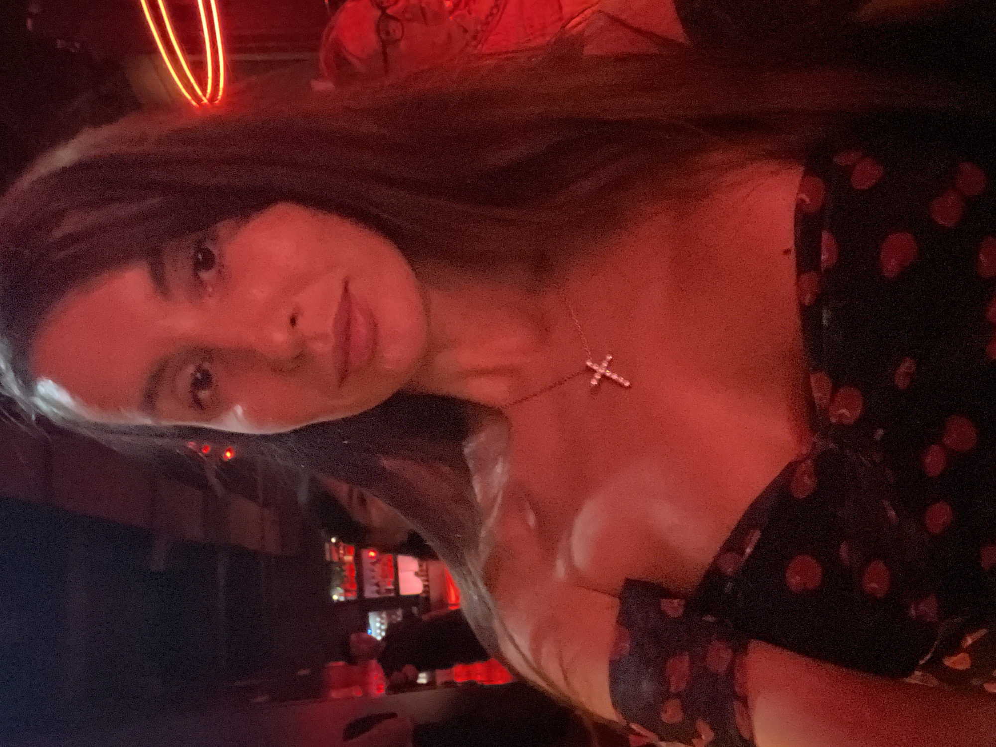 A woman is captured in an indoor setting with red lighting. She is wearing a dark-colored dress with red polka dots and a necklace with a cross pendant. The background includes a bar area with various bottles and a blurry figure of another person. There is a red neon light fixture above her head. There is no evidence or indication of this image being taken in North Kuta, Indonesia.