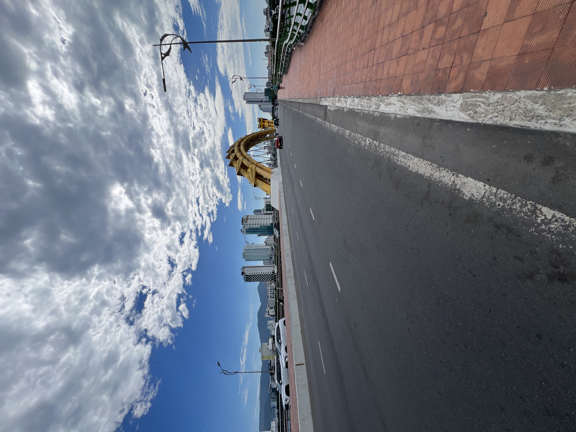 The image depicts the Dragon Bridge (Cầu Rồng) in Đà Nẵng, Vietnam, on a clear day with significant cloud cover. The perspective is from the dark asphalt roadway, looking forward towards the iconic golden dragon-shaped arch structure of the bridge. The roadway features white dashed lane markings. To the right, a grey concrete curb separates the roadway from a reddish-brown paver sidewalk. A green metal railing with a silver top rail runs along the right edge of the sidewalk, partially obscuring the river below.
On the roadway, a red sedan and a dark SUV are visible, driving towards the dragon structure. A single pedestrian walks away from the camera on the right-hand sidewalk. On the left side of the bridge, beyond a low white barrier, two white SUVs are parked, and a person on a scooter is further down the road. The cityscape of Đà Nẵng forms the background, characterized by multiple modern high-rise buildings. Distant, hazy mountains are visible on the horizon behind the buildings on the left. The sky features clear blue patches interspersed with dense, textured white and grey clouds. Two decorative streetlights, with black poles, fish-fin motifs, and horizontal light arms, are positioned on either side of the bridge span.