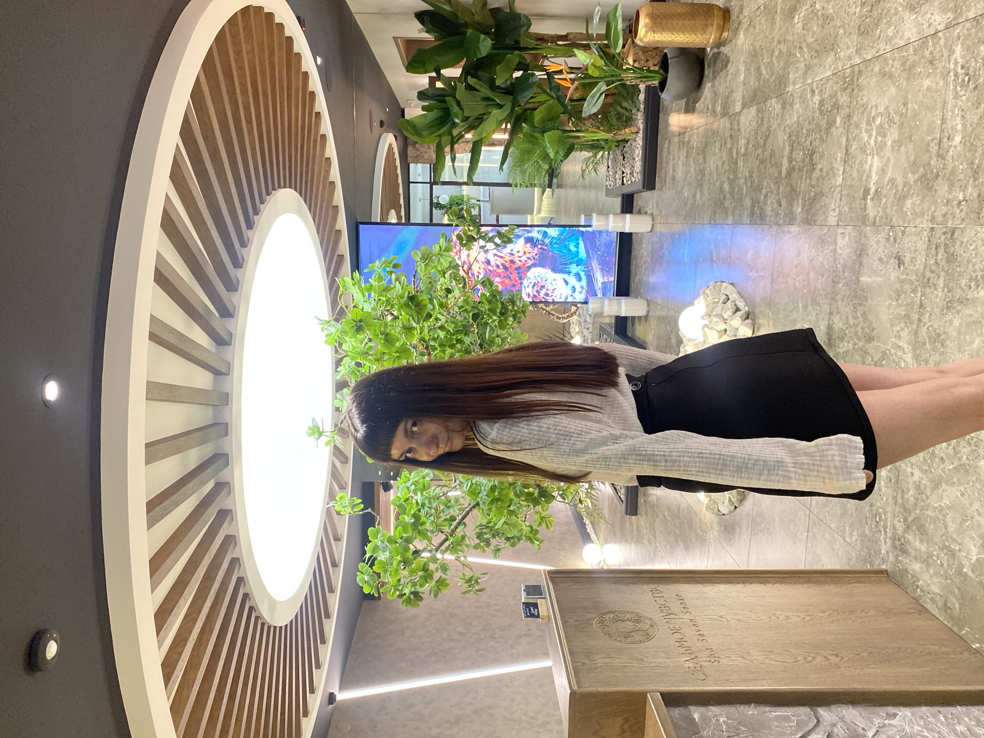 A young woman with long, dark hair and bangs stands in an indoor setting that appears to be a spa or reception area. She is wearing a white, long-sleeved, ribbed shirt and a short black skirt. She is looking over her shoulder at the camera with a slight smile.
The environment features a wooden reception desk with text in Cyrillic and English, a large vertical digital screen displaying a colorful image of what looks like a leopard and coral, and several potted plants. The ceiling has a circular design with wooden slats radiating outwards from a central illuminated panel. The floor is tiled with a grey, marbled pattern. The overall impression is modern and tranquil. The provided text on the reception desk, "СЕДЬМОЕ ЧУВСТВО Spa Seven Sense," indicates the location is likely a spa. Given the context, if this is in Votkinsk, Russia, it would be a commercial establishment within the city.
