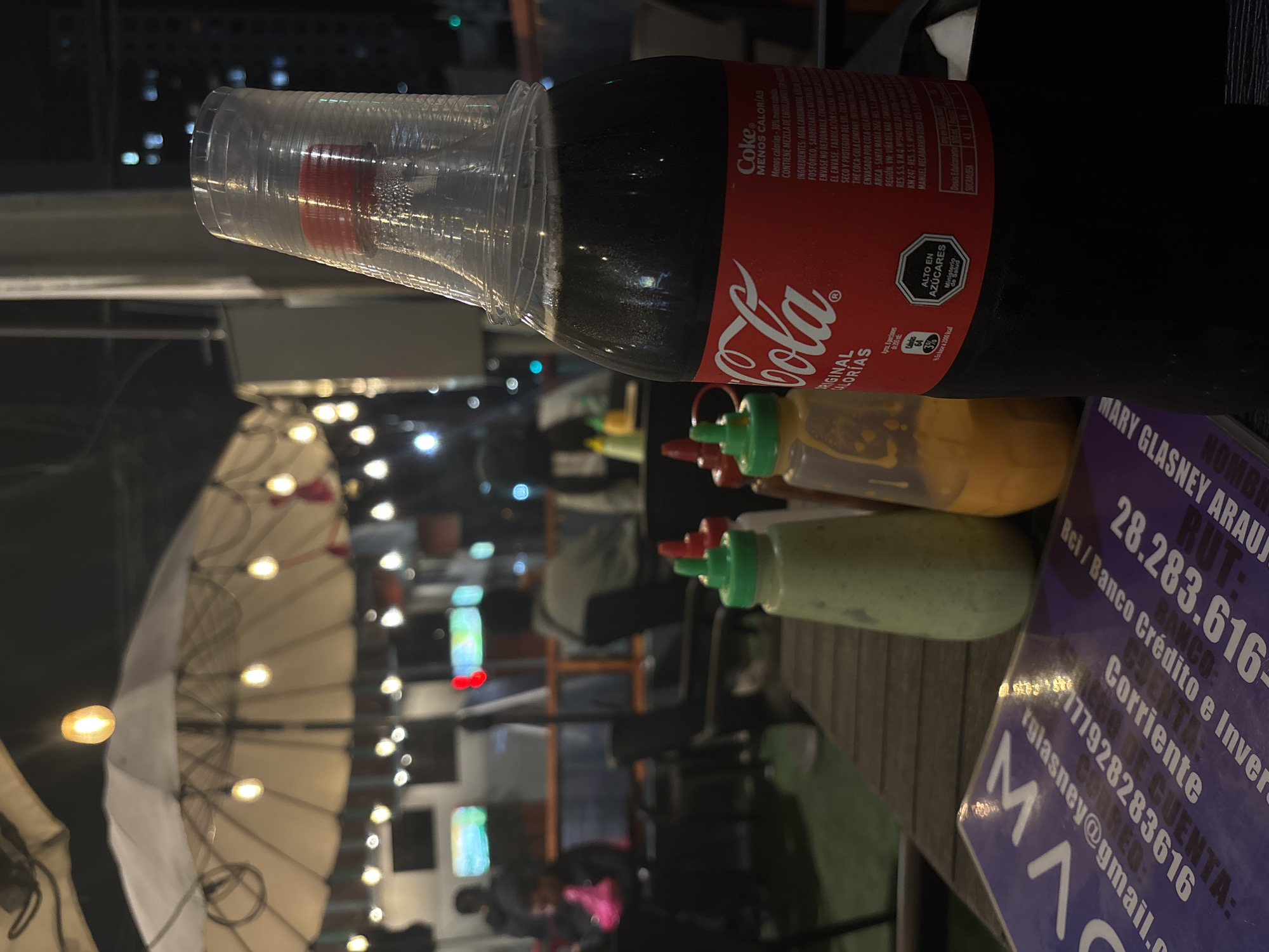 The image depicts a close-up of a plastic bottle of Coca-Cola, partially filled with a dark liquid, resting on a wooden table. The bottle has a red label with the Coca-Cola logo. Beside it are two condiment bottles, one with a green cap and a light green substance, and another with an orange-yellow substance. In the foreground, a portion of a purple booklet is visible with text that includes "MAR", "glasey@gmail.com", and various numbers and financial terms. In the background, an outdoor seating area is visible with tables, chairs, and string lights illuminating an umbrella. Several people are dimly lit in the background, some seated and others standing. The scene appears to be an outdoor dining establishment or bar at night, potentially in a city, suggested by the distant building lights. Based on the visible text on the booklet, which includes "RUT: 28.283.616-", a common format for Chilean identification numbers, and "iqlasey@gmail.com", which contains "iql" likely referring to Iquique, the location is suggested to be in the city of Iquique, Chile.