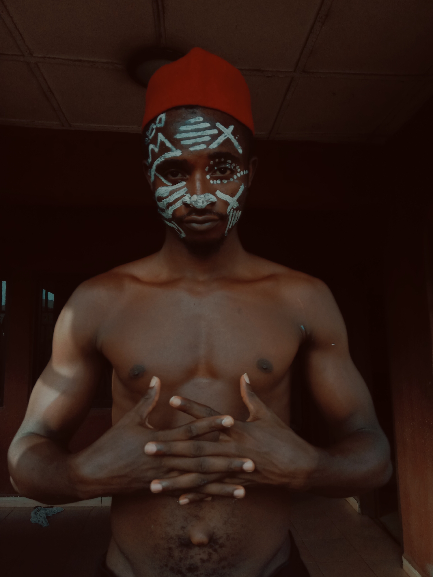 The image features a shirtless, dark-skinned man wearing a red cap, possibly Igbo traditional attire, and white face paint designs. He stands indoors, possibly within a building in Nkpor, Nigeria, as indicated by the prompt. He has dark eyes, and dark hair. He has his hands clasped and his fingers crossed over his stomach. The background shows dim lighting and a tiled ceiling. The environment appears domestic.