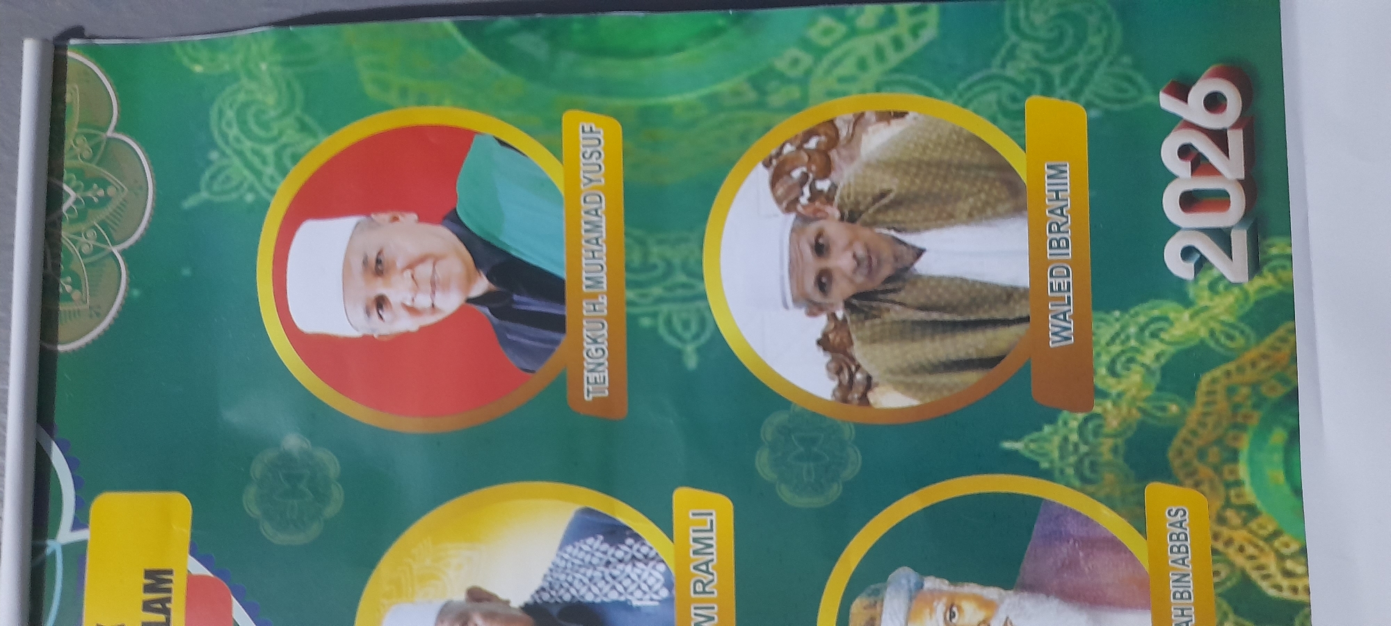 The image depicts a banner featuring portraits of Islamic religious figures. The banner is predominantly green with decorative gold accents. Two portraits are visible in the upper portion of the image. The first portrait, on the left, is of a man with a white cap, set against a red background within a gold circle. Next to this portrait, the name "TENGKU H. MUHAMAD YUSUF" is displayed in yellow text. The second portrait, to the right, is also of a man wearing a white cap, with a tan garment, framed within a gold circle. Adjacent to this portrait, the name "WALED IBRAHIM" is written in yellow text. To the right of the second portrait, the number "2026" is prominently displayed. The city of Buketteukuh, Indonesia, is not visible in the image.