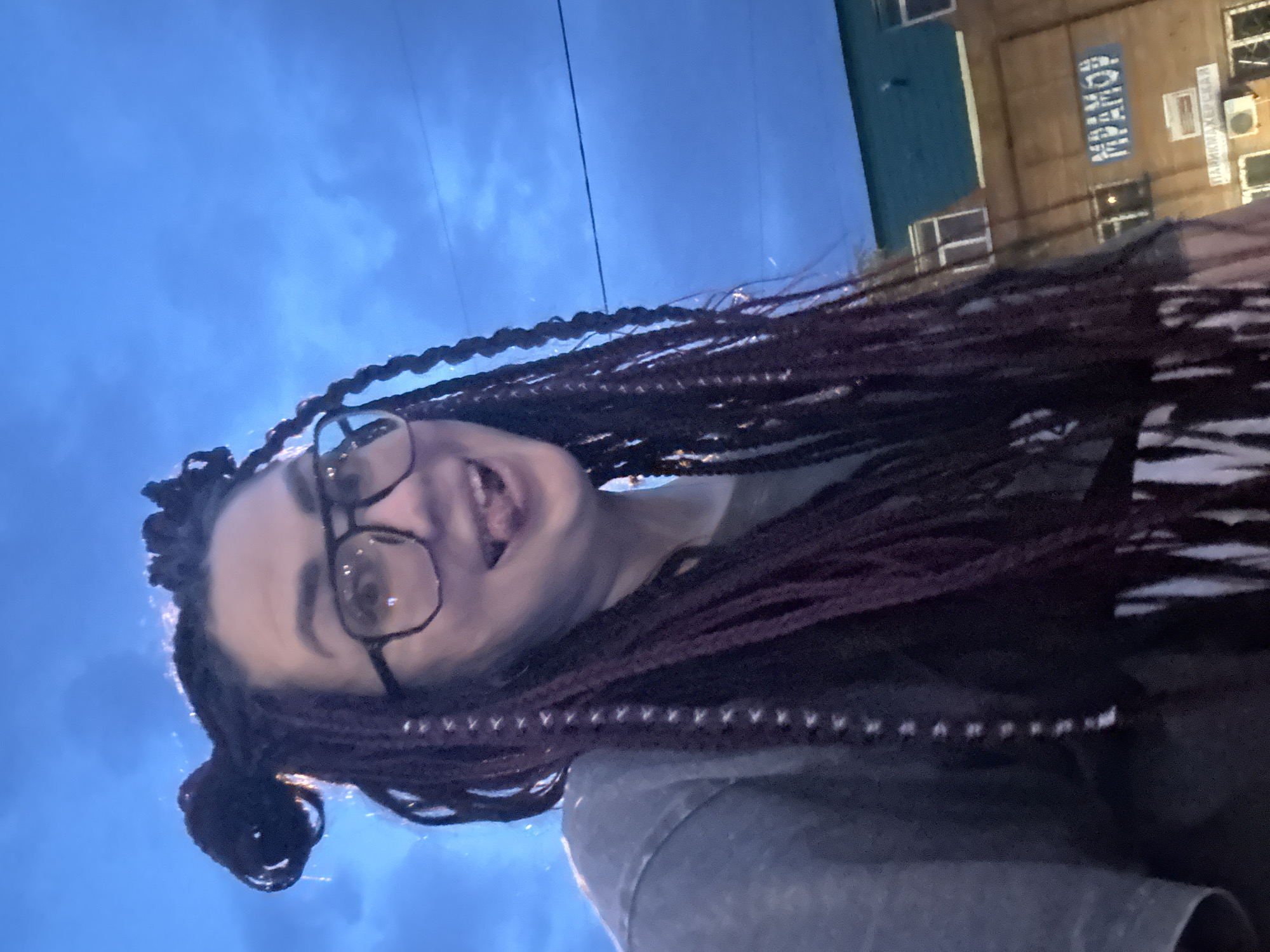 A young woman with dark, braided hair styled into two buns on top of her head and long braids hanging down, wearing glasses and a grey t-shirt, sticks her tongue out at the camera. She is positioned in the foreground, with a blurred blue sky and some clouds visible behind her. In the background, a light brown building with a teal roof is visible, with windows and air conditioning units. A sign on the building reads "MPAMOP" in blue lettering, and below it, another sign reads "ПАРИКМАХЕРСКАЯ" (which translates to "Hairdresser's"). The scene appears to be outdoors in Tolyatti, Russia, possibly in the evening given the lighting.