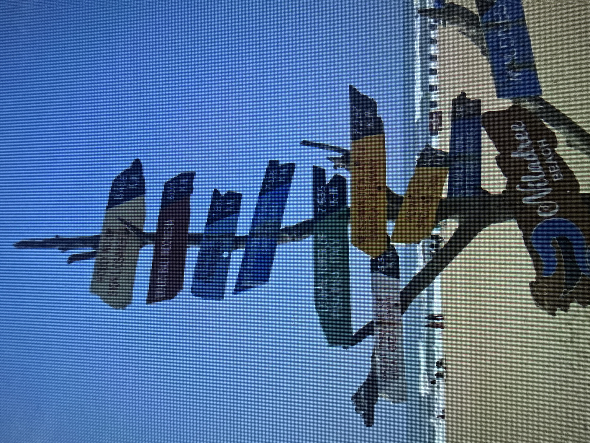 The image shows a directional signpost on a sandy beach. The signpost is made of weathered wood and has multiple signs pointing in different directions, indicating distances to various locations. Some of the legible signs include "HONEY MOON," "SINGAPORE," "BALI INDONESIA," "LEANING TOWER OF PISA ITALY," and "NEUSCHWANSTEIN CASTLE GERMANY." There are also some signs that are partially obscured or illegible. In the background, there is a clear blue sky, the ocean with waves, and a beach with a few people walking and sitting. Some beach chairs and umbrellas are visible in the distance. In the foreground, on the right side, a brown and blue sign with the words "Niladree BEACH" is visible. The overall scene depicts a tourist destination on a beach, with signs indicating travel distances to popular global landmarks.