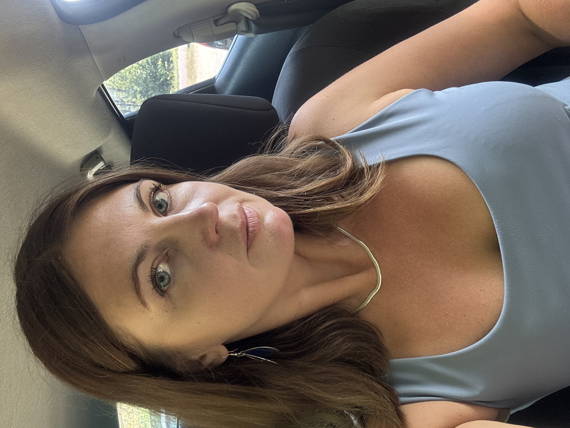 A woman with shoulder-length brown hair and light blue eyes is sitting in the passenger seat of a car. She is wearing a light blue tank top, a silver necklace, and a dark blue earring. The interior of the car is visible, including the headrest of the seat behind her and the ceiling of the car. Through the window, trees and foliage are visible. The location is in the municipality of Coteaux du Lizon, France.