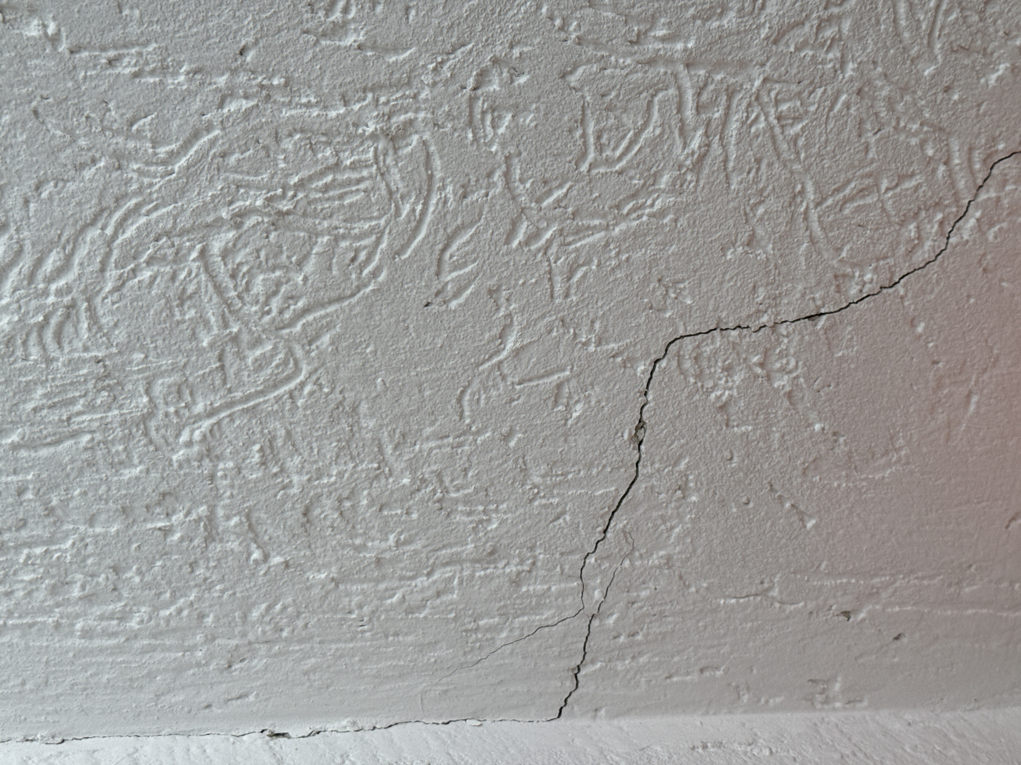 The image shows a close-up view of a textured white wall with a visible crack. The crack originates near the corner of the wall and extends diagonally across the surface. The wall appears to be an exterior surface, possibly of a building. The textured pattern on the wall is irregular, with raised lines and depressions. There are no people or other objects clearly visible in the frame. The lighting suggests it is daytime, with soft shadows indicating diffused light. The image does not contain any specific identifiable landmarks or information that would allow for pinpointing a location within Nizhny Novgorod, Russia.