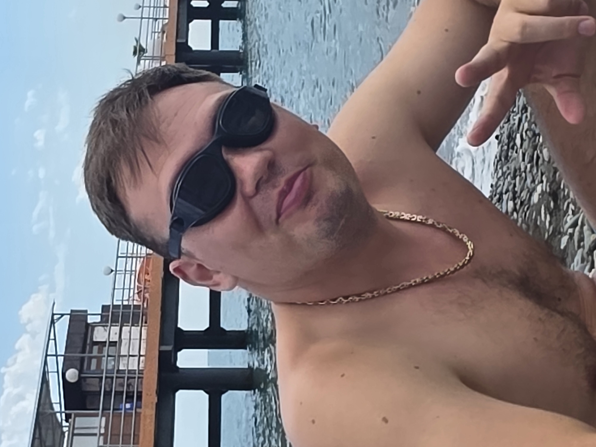 A man is shown from the chest up, seated on a pebble beach with the sea in the background. He is wearing black swimming goggles and a gold chain. Behind him, a pier extends into the water, with a building on stilts and a railing. The sky is partly cloudy. The location is identified as Sochi, Russia.