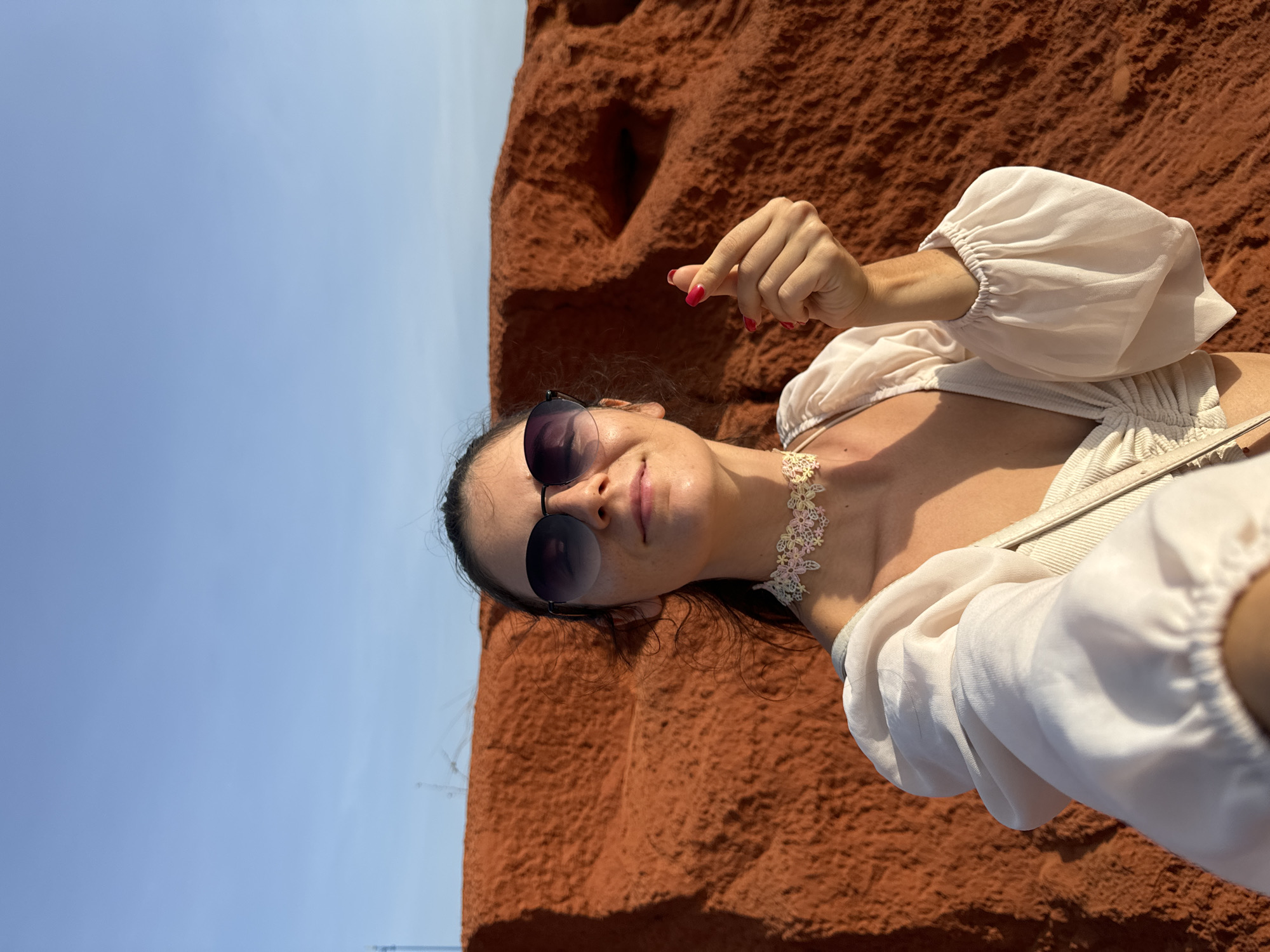A woman is taking a selfie in Mui Ne, Vietnam, in front of the red sand dunes. She is wearing sunglasses, a white top with puffed sleeves, and a floral choker. Her right hand is extended, forming a heart shape with her thumb and index finger. The background consists of the distinct red sand formations characteristic of the area, with a clear blue sky above.