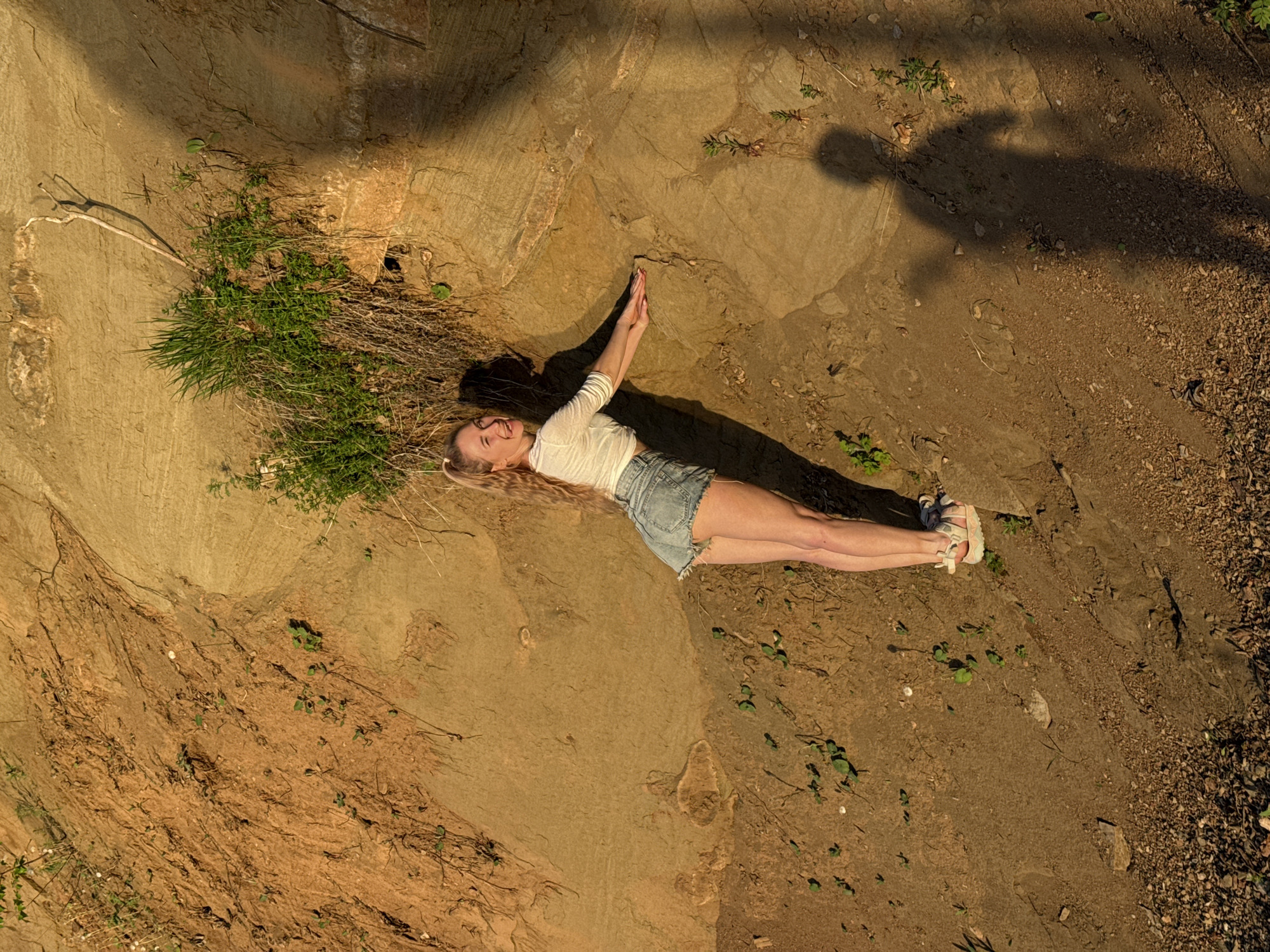 A bird's-eye view photograph captures a young woman lying on a rugged, earth-toned landscape. The setting appears to be outdoors in the city of Chaikovsky, Russia.
The woman, fair-skinned with blonde hair pulled back, is clad in a white long-sleeved shirt and denim shorts, and sandals. She is lying prone, with her arms extended above her head, her palms pressed together. Her shadow is visible behind her, and a separate human shadow falls across the top-right of the frame, indicating the photographer.
The terrain beneath her is uneven, displaying a mix of rock, dirt, and sparse vegetation, including some grass and small green plants. The color palette is dominated by browns, tans, and muted greens, suggesting a dry or rocky environment.