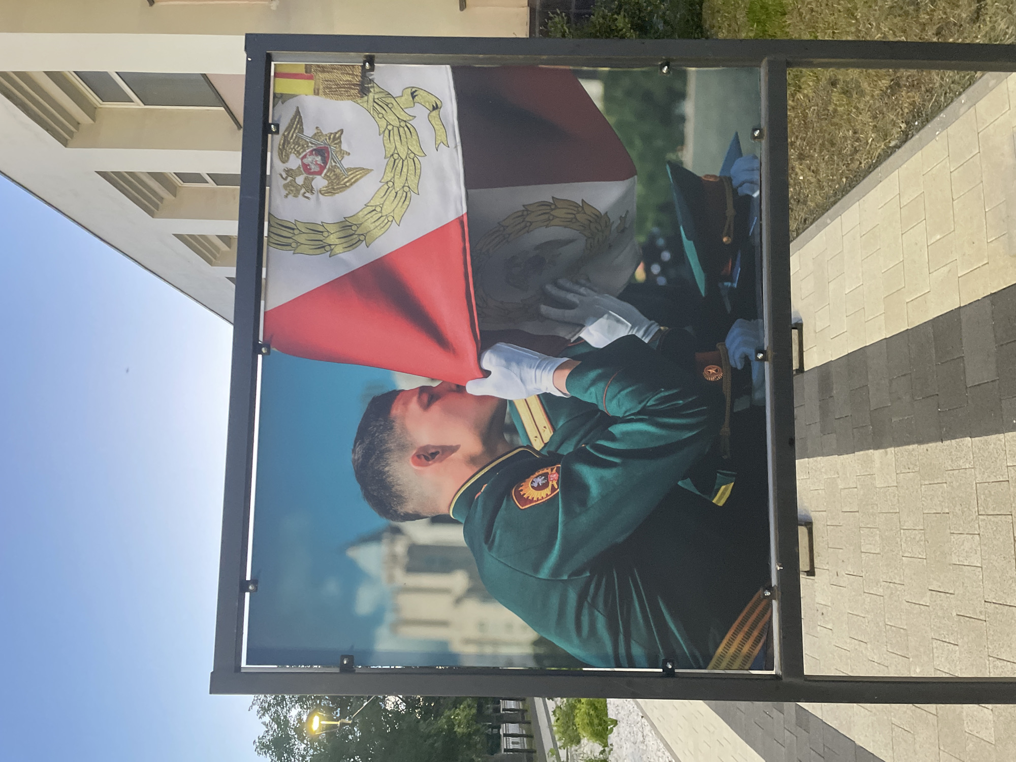 The image shows a large outdoor banner displaying a photograph of a young man in a dark green military uniform kissing a flag. The man is wearing white gloves and a cap. The flag is red and white with a circular emblem featuring a double-headed eagle and a laurel wreath. The banner is framed by a black metal stand. In the background, a building with cream-colored walls and windows is visible, along with trees and a street lamp. The ground in front of the banner is paved with light-colored rectangular tiles. The scene appears to be an outdoor public space in Vologda, Russia, possibly near a government building or monument. The lighting suggests it is daytime, with a clear blue sky.
