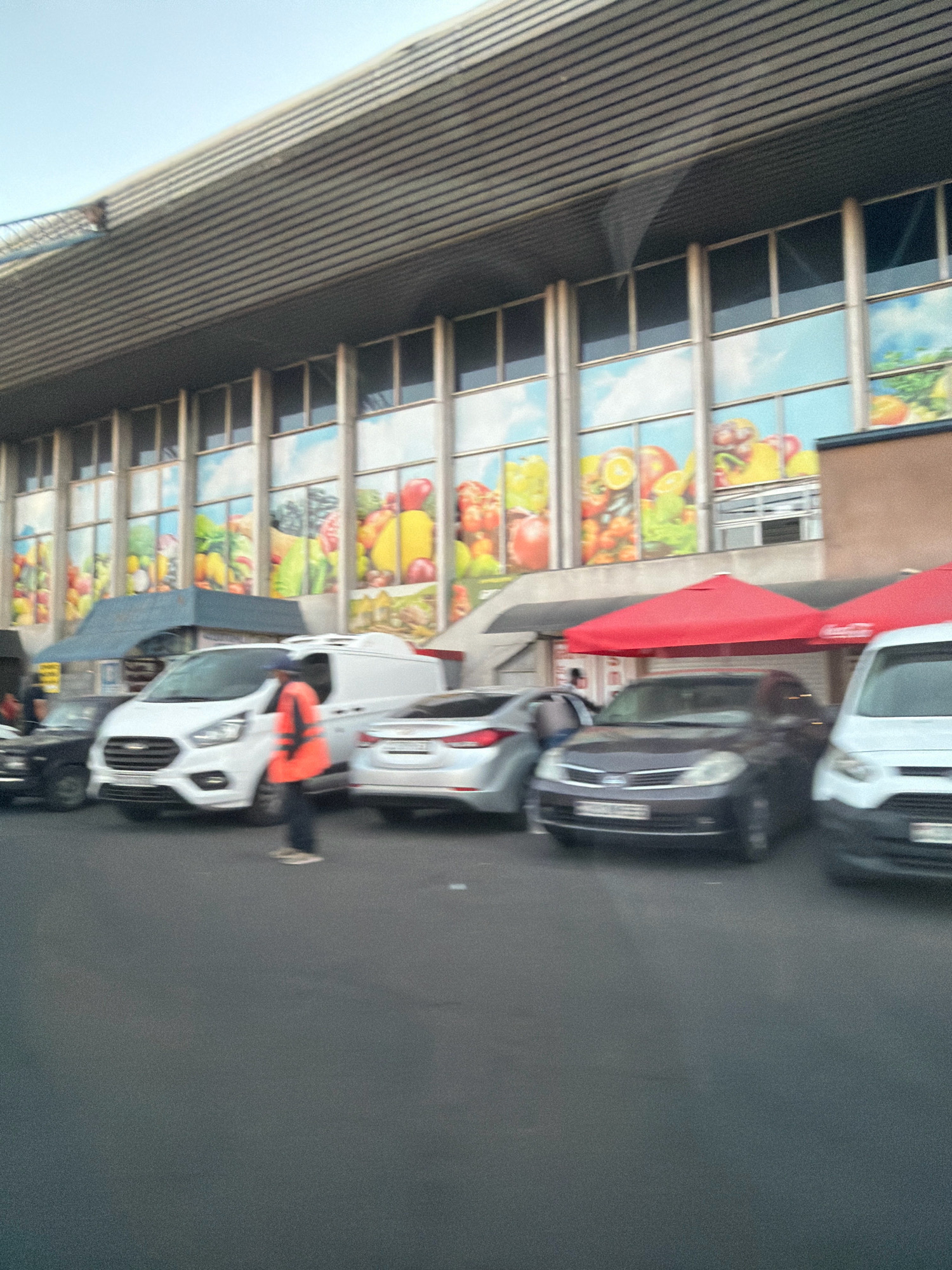 The image captures a street scene in Yerevan, Armenia, in front of a building with large, colorful murals depicting fruits and vegetables. The building's facade features a modern design with horizontal metallic louvers across the top and multiple vertical windows. In the foreground, several vehicles are parked, including a white van, a silver sedan, and a dark sedan. A person wearing an orange vest and dark pants is standing on the asphalt, appearing to direct traffic or perform some task. Red umbrellas are set up near the building, suggesting an outdoor market or food stalls. The overall impression is a bustling urban environment, possibly a market or commercial area, with a blend of modern architecture and vibrant street life.