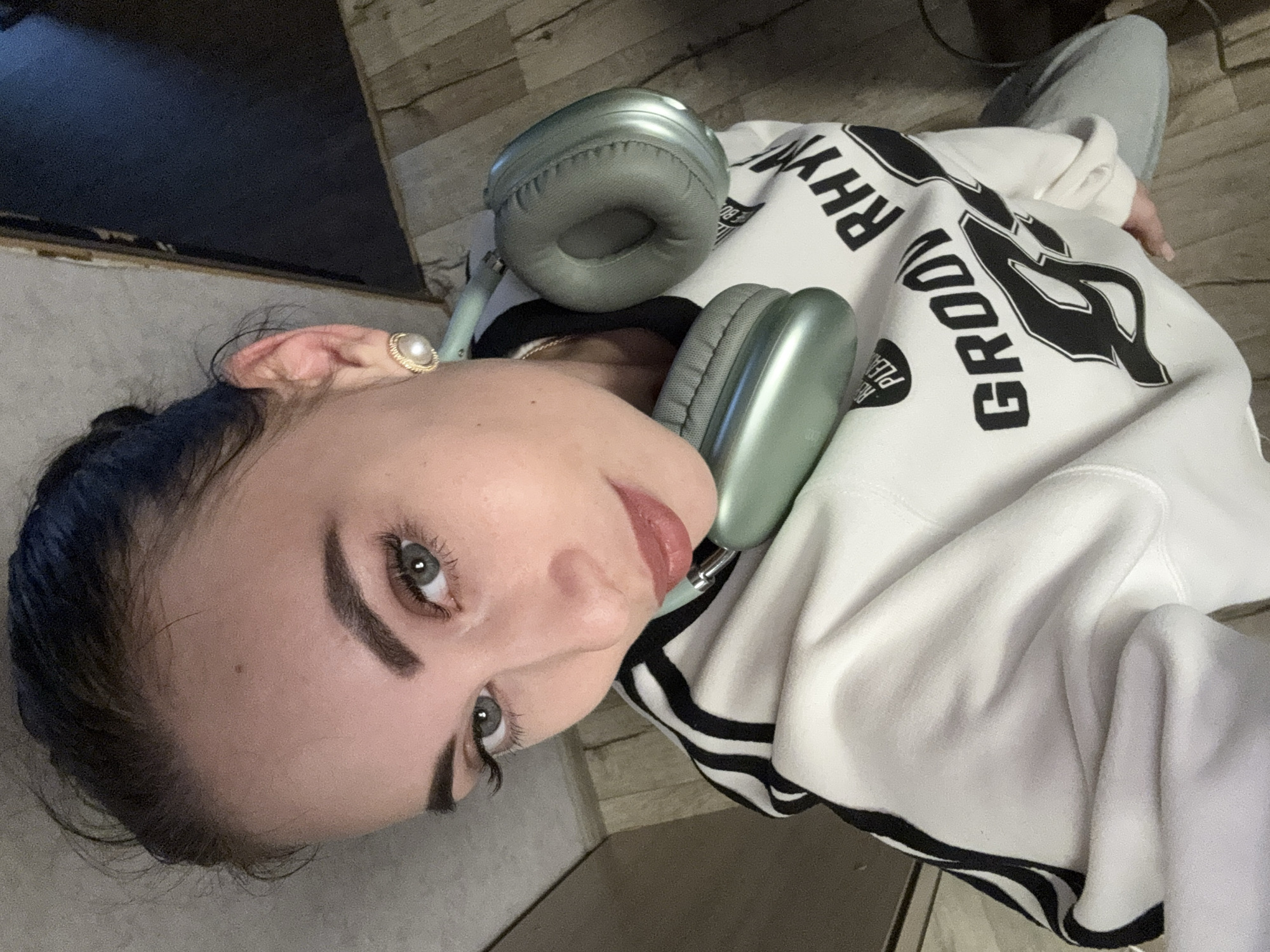 A woman is seated indoors, wearing a white hoodie with black stripes and the word "GROOVY" printed in large black letters on the front, along with the words "RHYTHM" and "PLEASE". She has light skin, dark eyebrows, and bluish-dark hair pulled back. Gray-green headphones rest around her neck. She is wearing a gold earring with a pearl. The background includes a dark gray surface and light-colored flooring. The location is specified as Chaikovsky, Russia.